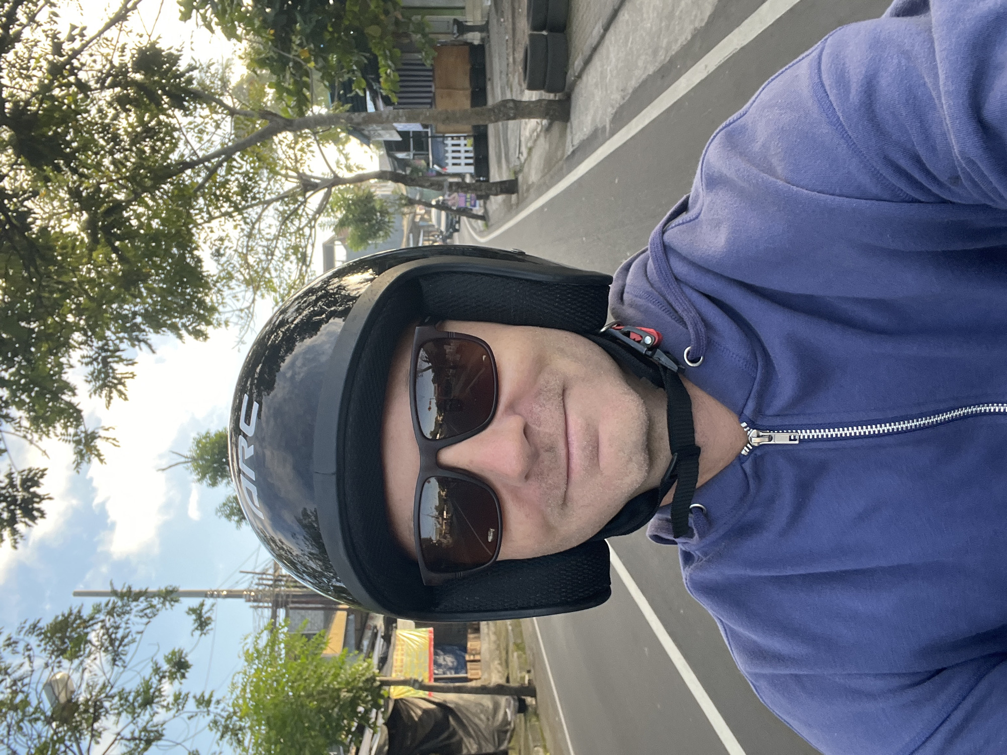 The image shows a man wearing a black helmet and sunglasses, riding a motorcycle on a street in Abiansemal, Indonesia. The man is smiling and looking at the camera. He is wearing a blue hoodie. The street is paved and has white lines marking the lanes. There are trees and buildings on both sides of the street. The sky is blue with some clouds. There are other vehicles visible in the background, including a car and a motorcycle. The scene appears to be during the daytime, with the sun casting shadows.