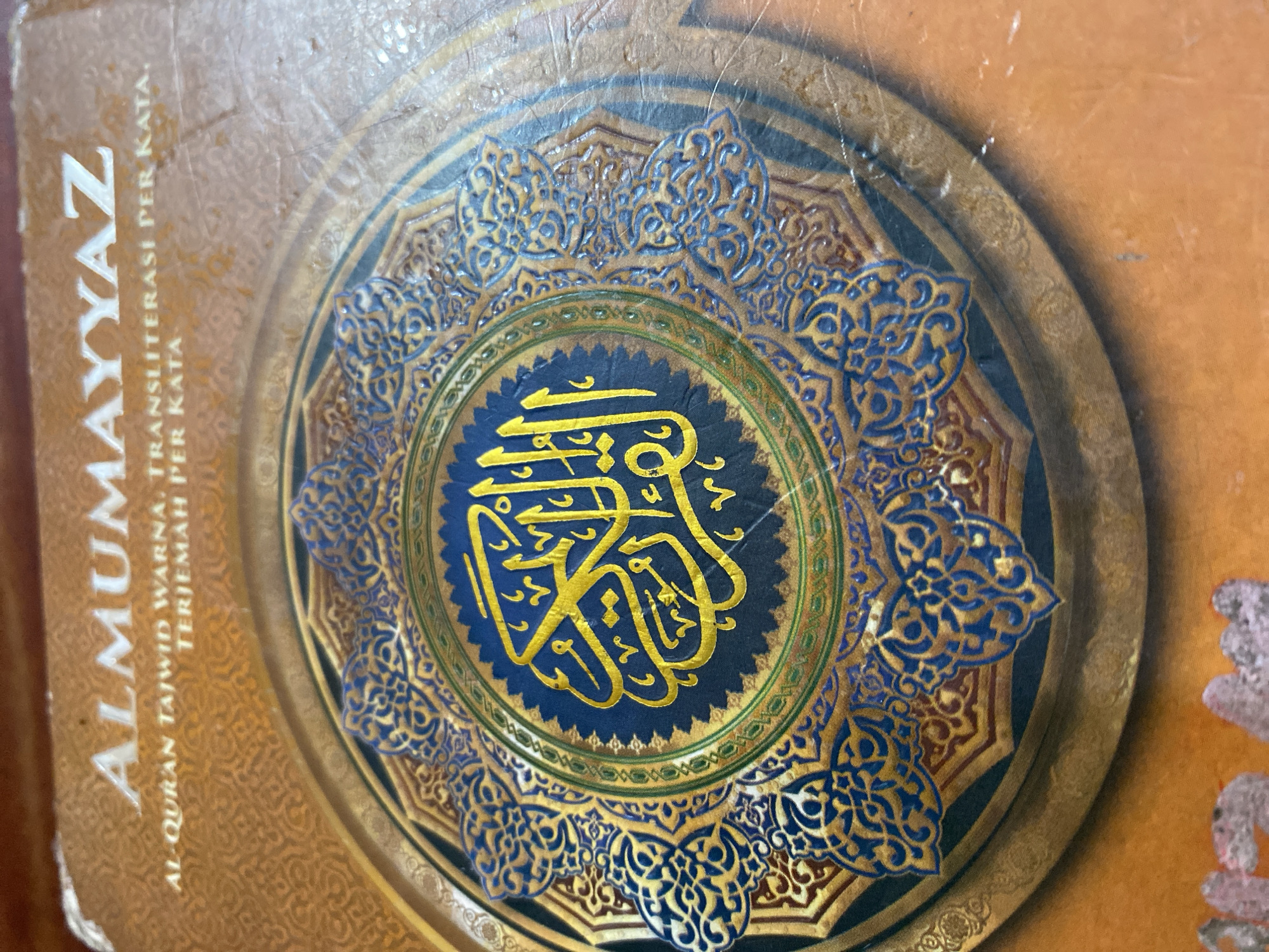 The image shows the cover of a book titled "AL-MUMAYYAZ". The book cover features intricate Islamic geometric patterns in gold and blue on a brown background. At the center of the cover is a circular design with Arabic calligraphy in gold. The text "AL-QURAN TAJWID WARNA, TRANSLITERASI PER KATA, TERJEMAH PER KATA" is printed above the circular design. The book appears to be an edition of the Quran with color-coded Tajwid, word-for-word transliteration, and translation. The image is a close-up shot of the book cover and does not contain any information about its location within the city of Ta', Indonesia, or any people or actions.