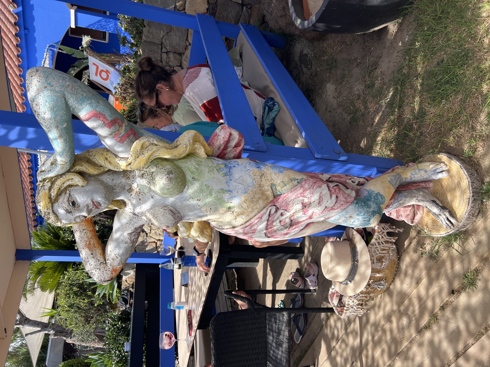 A statue of a woman with a painted, weathered finish stands in the foreground, positioned on a wooden base. The statue is adorned with colorful patterns, resembling a map or abstract designs. To the right of the statue, a bright blue wooden bench with cushions is occupied by two women. One woman, on the right, wears a striped red and white top and sunglasses, her hair is tied up. The other woman, to her left, is partially visible, wearing a light green top. In the background, a wooden table is set with drinks, glasses, and phones. Beyond the table, lush green vegetation and a building with a red-tiled roof are visible. The scene appears to be an outdoor seating area, likely at a restaurant or cafe in Tarifa, Spain, given the style of the architecture and vegetation.