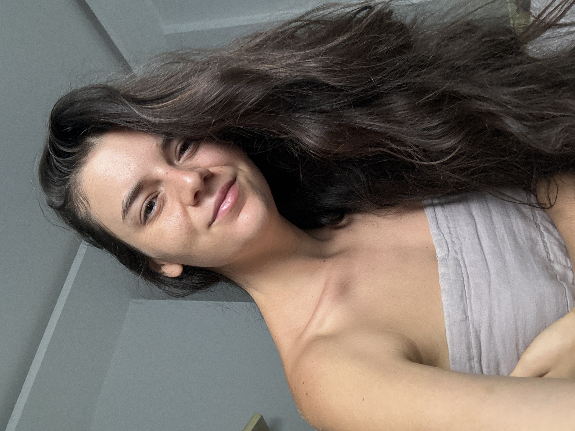 A young woman with long, wavy dark hair smiles at the camera. She has fair skin, brown eyes, and freckles on her face. She is wearing a light-colored tank top. The background consists of a gray wall and a small section of what appears to be a doorframe in the lower left corner. The location is not discernible from the image.