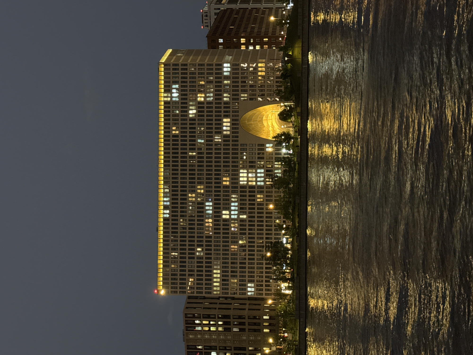 Captured at night, the image features a tall, illuminated building with a prominent arched structure in the middle, possibly located in an urban area with a body of water nearby. The building is adorned with numerous windows, many of which are lit, giving the structure a grid-like appearance. The edge of the building is illuminated with a continuous line of light. To its side, a darker building is partially visible. The water in the foreground reflects the lights from the building, creating a shimmering effect. The sky is dark, suggesting it is either late evening or night.