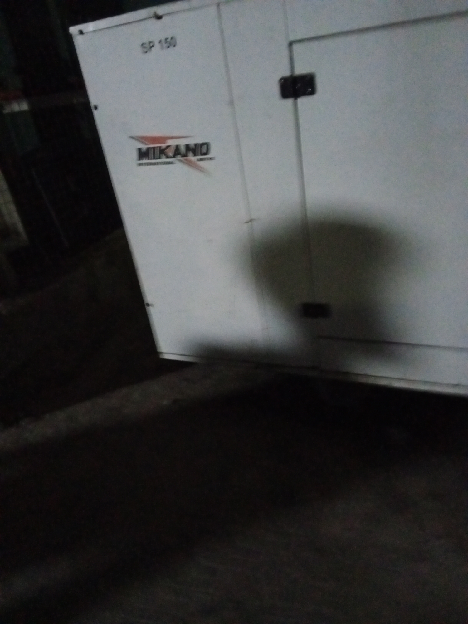 A white Mikano generator, model SP 150, sits on a dark surface, possibly concrete, inside a poorly lit building in Ado, Nigeria. The lighting is dim and casting shadows. The generator's logo is visible on its side. The surrounding environment is dark and indistinct, with other objects out of focus in the background. The scene appears to be indoors.