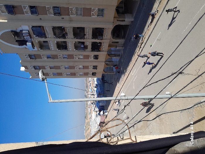 A high-angle shot reveals a street scene in front of a tall building. Several people are visible on the street. A white line runs along the edge of the road near the building. Power lines crisscross the lower portion of the image. The sky is bright blue. A portion of a roof is visible in the foreground.