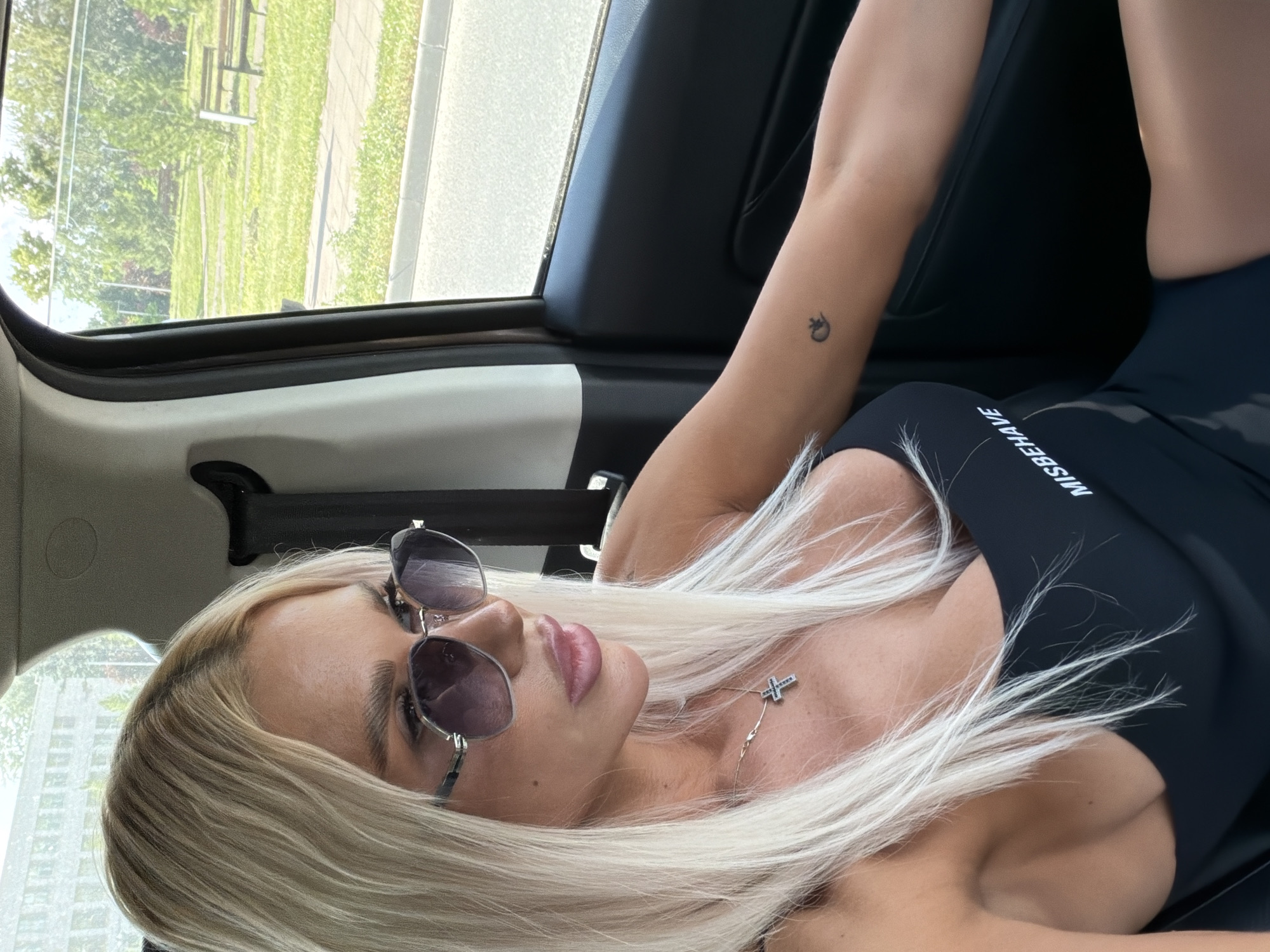 A blonde woman wearing sunglasses and a black top is sitting in a car. The car's interior is visible, along with a window looking out onto a green park with benches. The woman has a cross necklace and a small tattoo on her arm. The scene appears to be in Kazan, Russia.