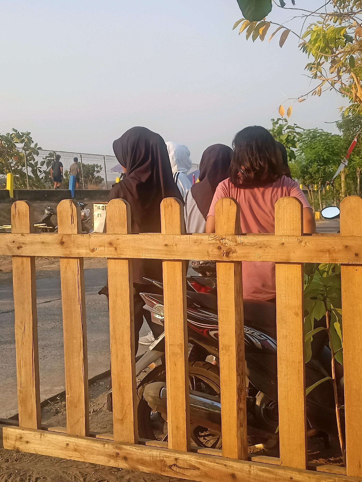 In the city of Ketro, Indonesia, a wooden fence forms the foreground of the image. Beyond the fence, three individuals are partially visible, all facing away from the camera. The person on the left wears a black hijab. The person in the middle wears a white hooded garment. The person on the right has dark, shoulder-length hair and wears a pink shirt. They appear to be standing next to a parked motorcycle, parts of which are visible through the fence. In the background, two men are walking away from the camera, one carrying a blue object. The environment suggests an outdoor setting with trees and a light-colored sky, possibly during sunrise or sunset.