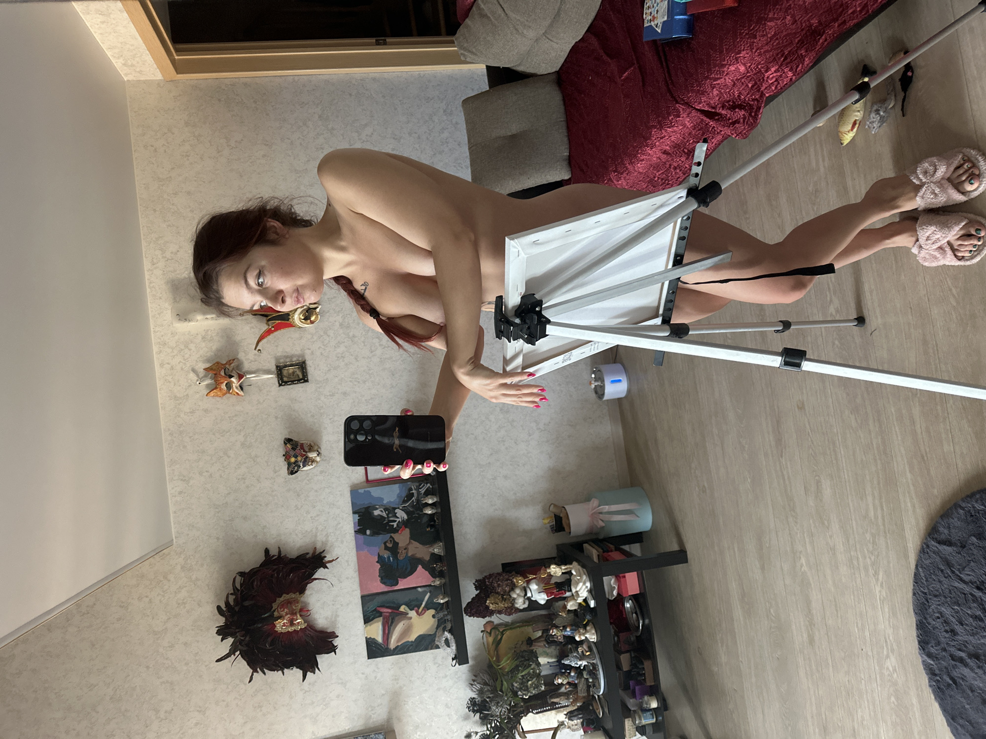 The image features a young woman standing partially nude in what appears to be an indoor room. She has reddish-brown hair styled in a braid and is wearing pink fluffy slippers. Her left hand holds a black smartphone, seemingly aimed at herself, while her right hand rests on her thigh. She is positioned next to a silver easel holding a blank white canvas, suggesting an artistic activity or pose. Her fingernails are painted pink, and a small tattoo is visible on her left shoulder/chest area. She looks directly at the camera with a neutral to slightly serious expression.
The room has light-colored, patterned wallpaper and a light wood laminate floor. In the background, there's a sofa or bed with red and grey upholstery, and a blue box rests on it. Various decorative items adorn the wall, including several masks (some resembling Venetian styles, one made of feathers) and a small framed picture. To the right, a black shelving unit displays numerous small objects, including bottles, jars, and figurines. A round, grey fluffy rug lies on the floor. Near the easel, a small blue and white cylindrical device is visible. No discernible text is visible within the image.