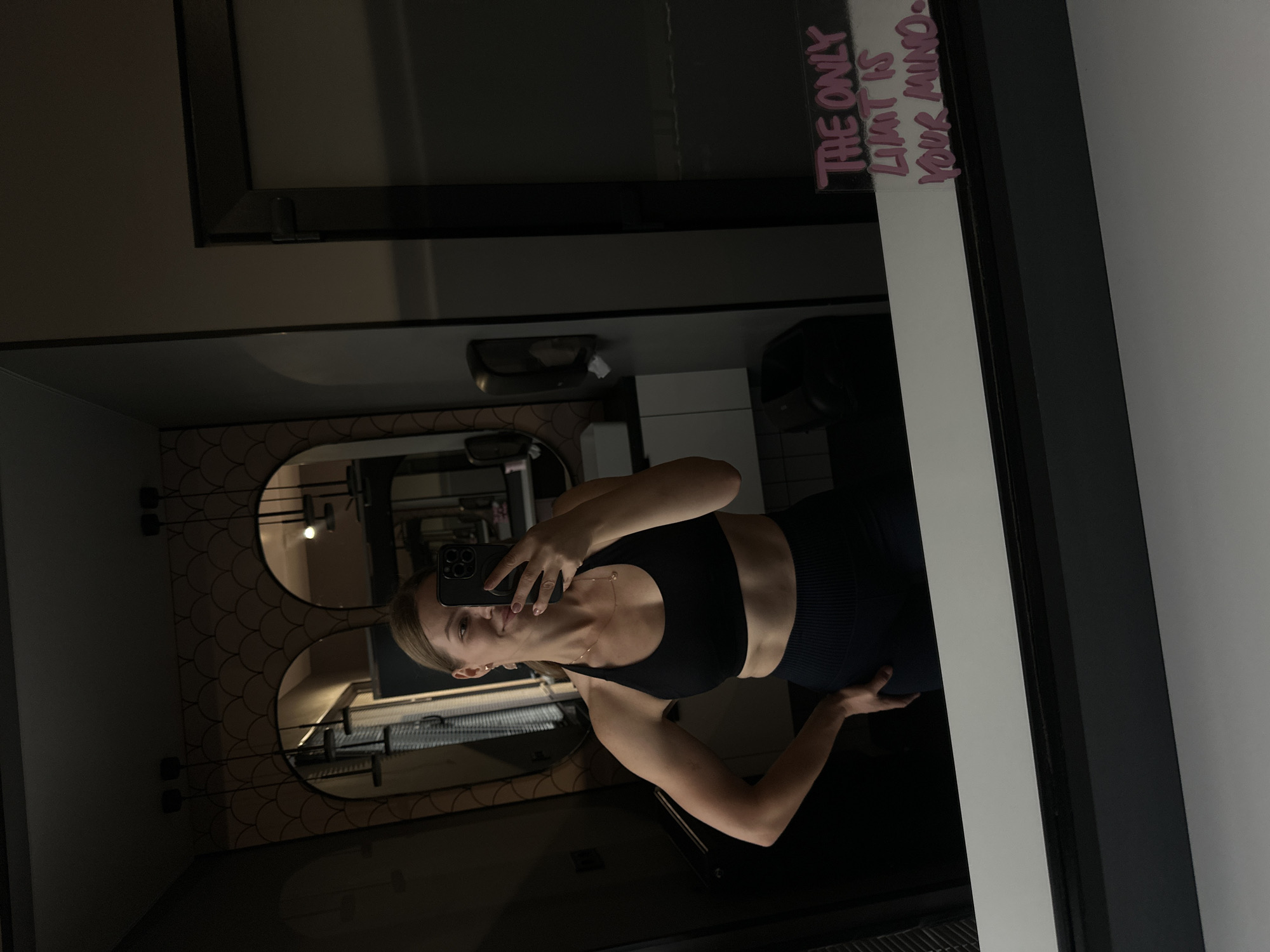 A young woman with blonde hair tied back, wearing a black sports bra and black leggings, takes a selfie in a mirror. She is standing in what appears to be a locker room or bathroom, reflected in a large mirror. Behind her, the reflection shows two arched mirrors with decorative tiling and light fixtures. On the right side of the main mirror, there is graffiti that reads "THE ONLY LIMIT IS YOUR MIND". The woman is smiling and has her left hand on her hip, holding a phone in her right hand. The lighting is dim. The location is not specified as Ternopil, Ukraine, or any other specific city.