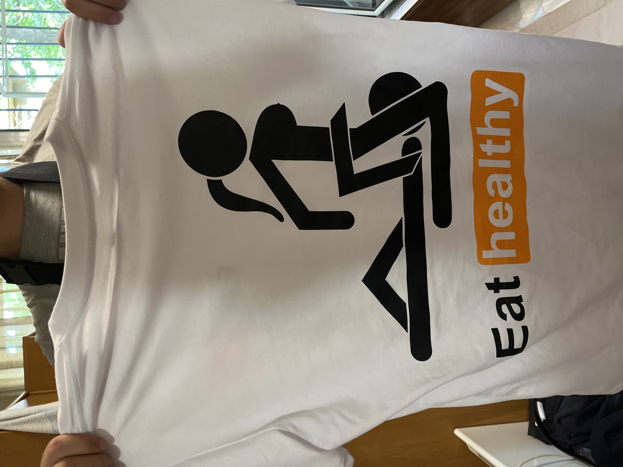 A person, whose hands and forearms are visible, holds up a white short-sleeved t-shirt. The individual is wearing a grey t-shirt and a dark strap across their right shoulder. The t-shirt features a large black silhouette graphic depicting two stick figures in a sexual position, with one female figure (distinguished by a ponytail and breasts) positioned on top of a male figure. Below this graphic, the text "Eat healthy" is printed. "Eat" is in black uppercase letters, and "healthy" is in white uppercase letters within an orange rectangular box, stylized to resemble the logo of a known adult entertainment website. The background on the left consists of light brown wooden furniture, with a white object and crumpled dark fabric below it. On the right, a window with visible green foliage and a dark metal railing outside indicates an outdoor view. No specific visual features are present in the image to confirm the location as Plovdiv, Bulgaria.