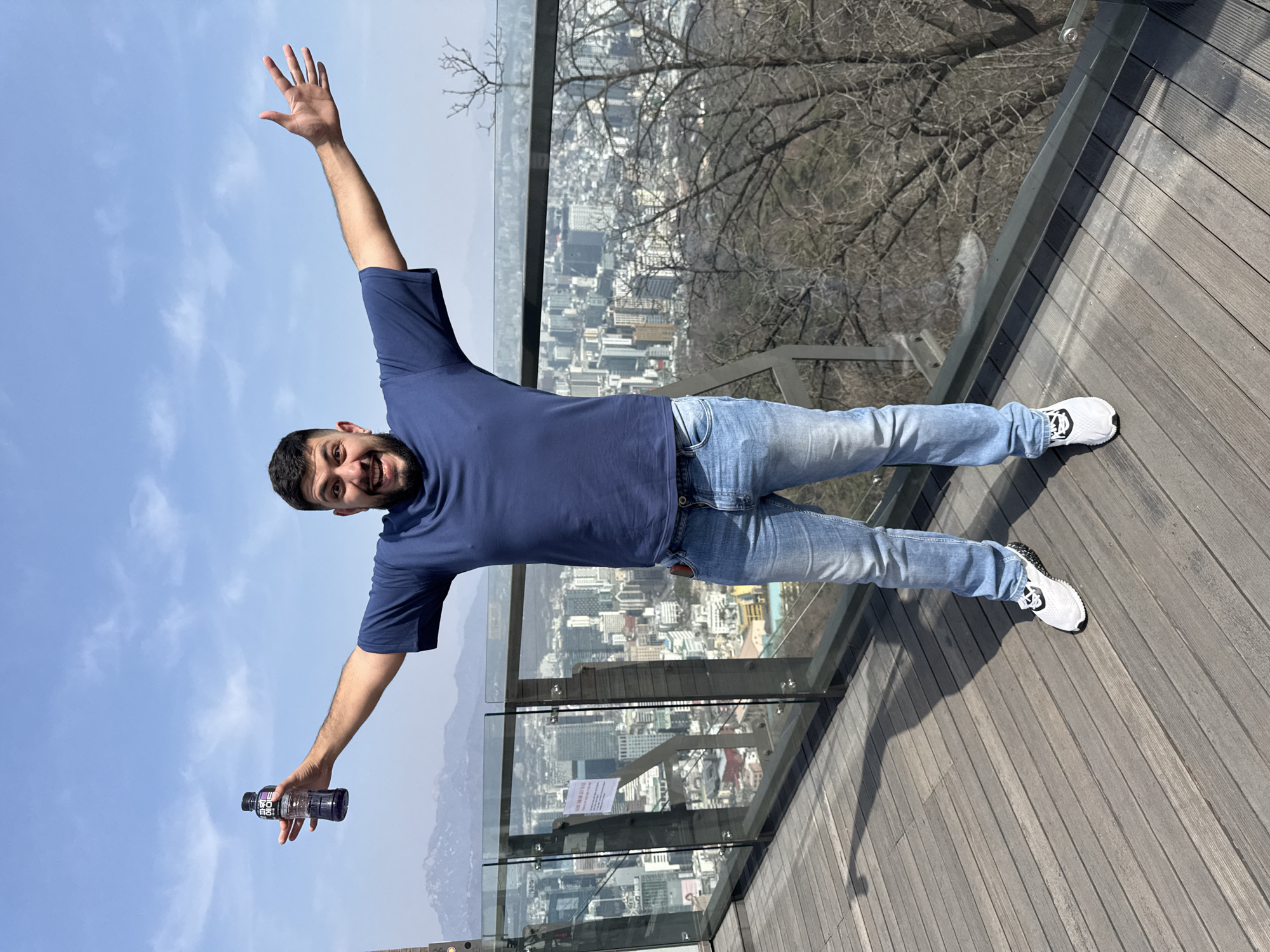 An adult male subject is standing centrally on a wooden slatted observation deck at an elevated viewpoint overlooking the city of Seoul, South Korea, likely from Namsan Mountain. The subject, with dark hair and a beard, is wearing a dark blue short-sleeved t-shirt, light blue denim jeans, and white athletic shoes with black soles. He is facing the camera directly, smiling broadly with visible teeth, and has both arms outstretched horizontally. His left hand holds a clear plastic bottle containing a purple liquid, identifiable as "Powerade" by Korean text on its label.
Behind the subject, a transparent glass and dark metal railing separates the deck from the urban landscape. A white sign with Korean text is affixed to one of the glass panels. Beyond the railing, a sprawling cityscape of Seoul is visible, featuring numerous high-rise buildings and urban structures extending to the horizon. Bare, leafless trees are visible on the hillside below and to the right of the railing. Distant mountains with a hazy outline appear on the far left horizon. The sky is clear blue with minimal wispy clouds. Direct sunlight is evident from the distinct shadows cast on the wooden deck.
