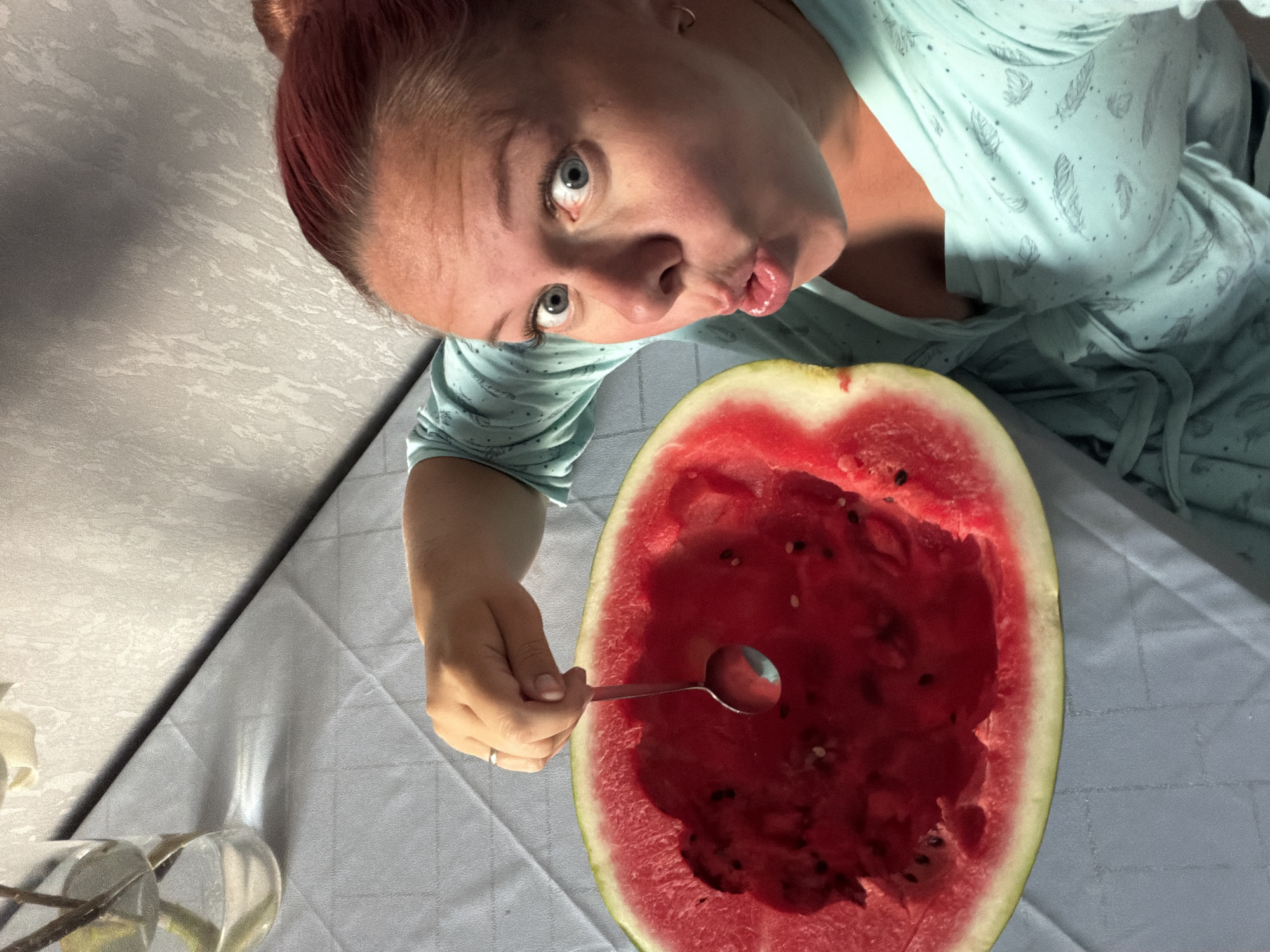 A woman with reddish-brown hair pulled up in a bun, wearing a light blue robe with a feather pattern, holds a spoon over a hollowed-out watermelon. Her eyes are wide and her mouth is open in a surprised expression. The watermelon is on a table covered with a light blue checkered tablecloth. In the background, a vase with branches and leaves is visible on the table, along with a textured wallpapered wall. The scene appears to be indoors, possibly in a home setting. The image does not provide specific details about the location in the city of Chaikovsky, Russia.