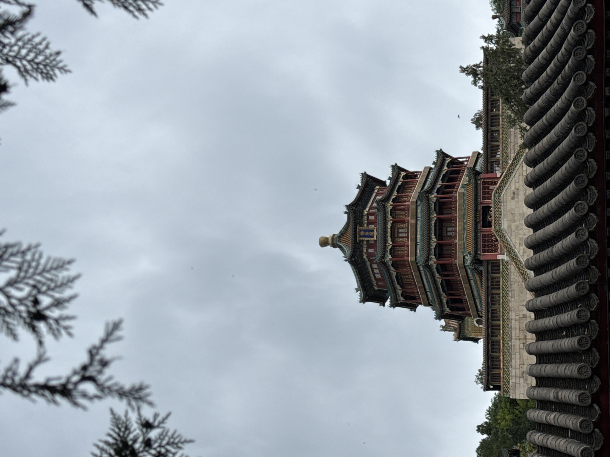 The image displays a towering, multi-tiered Chinese pagoda-style structure, specifically the Tower of Buddhist Incense (Fo Xiang Ge) at the Summer Palace, located in the Haidian District, Beijing, China. The structure is predominantly red and green with ornate detailing, featuring multiple eaves and a golden finial at its apex. It rises from an elevated stone platform, revealing other traditional buildings and lush green trees around its base. In the immediate foreground at the bottom, a row of dark grey traditional Chinese roof tiles, each with a decorative circular end, partially frames the lower part of the scene, indicating another structure closer to the viewer. The upper portion of the image, including the top of the pagoda, is set against a uniformly overcast, light grey sky, with blurred dark green coniferous foliage framing the top left and top right corners. Several tiny dark specks, likely birds, are visible against the sky. No individuals are present in the frame.