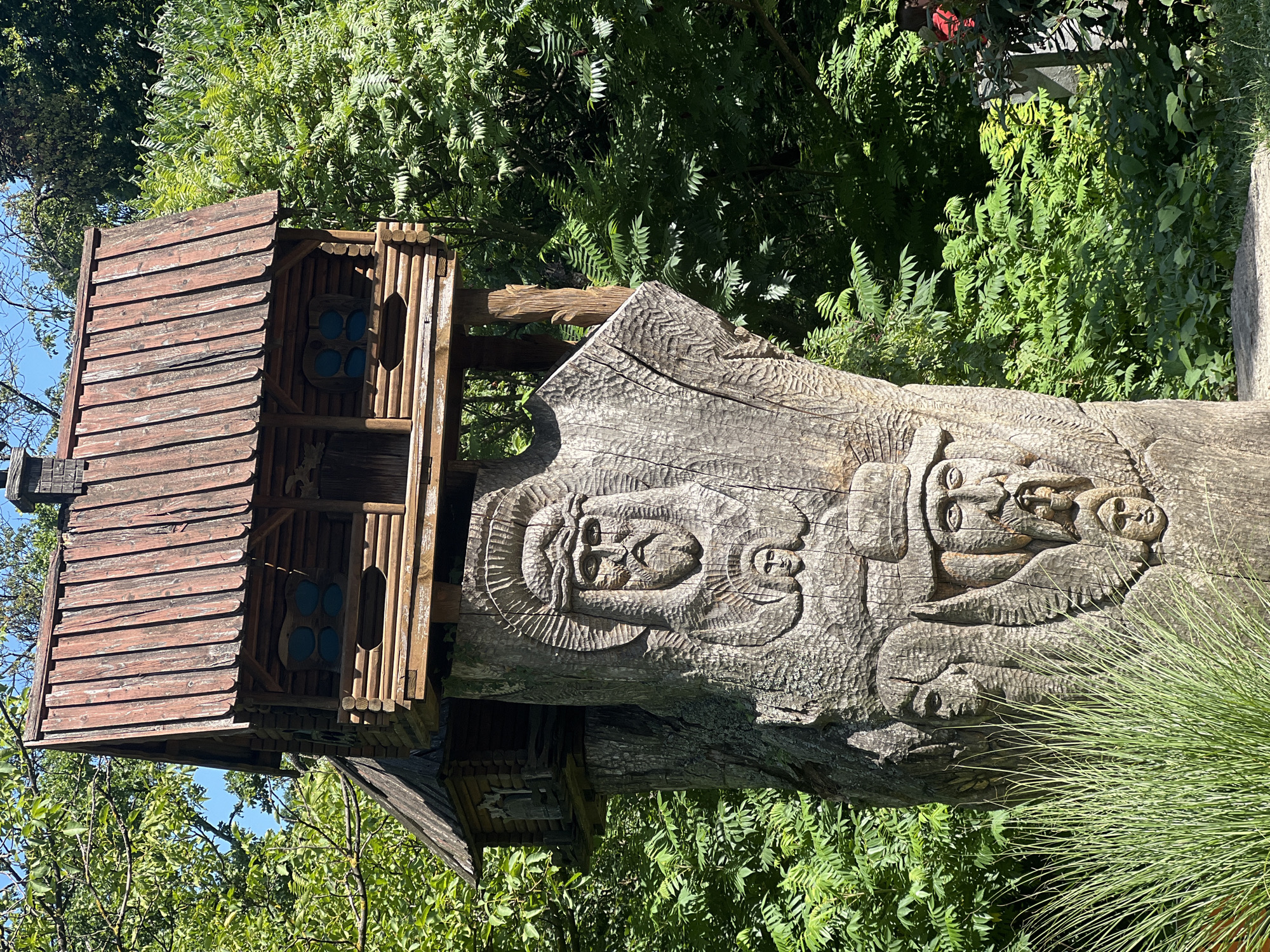 This is a medium shot, eye-level, vertical photograph of a large wooden sculpture in an outdoor setting. The sculpture is a tree trunk carved with multiple faces. The primary carving depicts a bearded face with a halo, likely representing a religious figure, with a smaller face positioned below it. To the right of this, another carving features a face wearing a hat, and below that, a very small face. To the left, another carved face is partially visible. Atop the wooden trunk, a miniature wooden house with a shingled roof has been constructed, appearing to be a birdhouse or decorative structure. The sculpture is surrounded by lush green foliage, suggesting a park or wooded area. The lighting indicates a bright, sunny day with shadows cast by the sun. The location is identified as Karpaty, Ukraine. There are no people actively engaged in actions within the frame, though a glimpse of a person in a red shirt is visible in the far right background. The carving itself is weathered, showing the texture and grain of the wood.