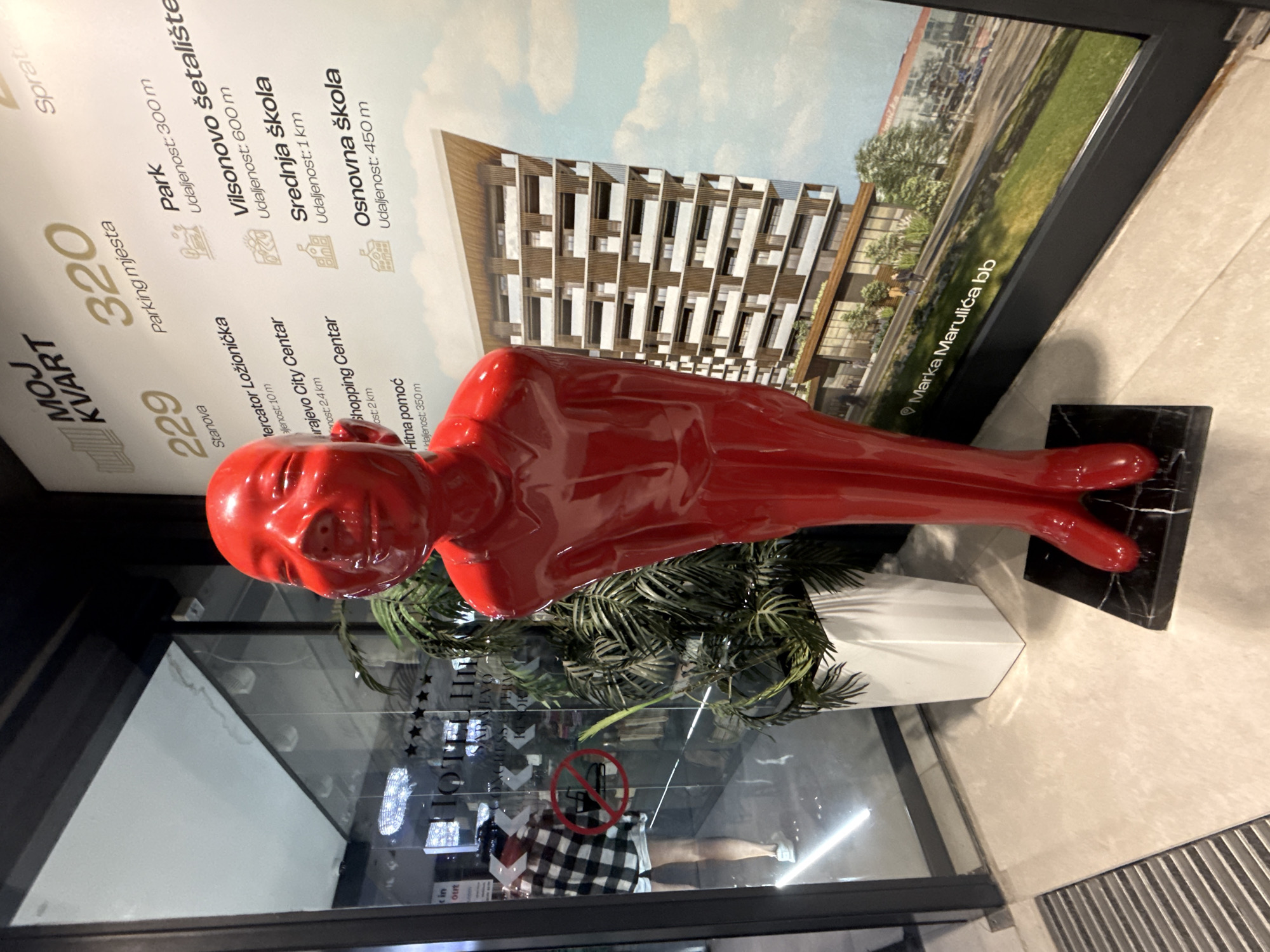 A red, glossy, humanoid sculpture with a child-like, smiling face stands in the foreground. It is positioned on a black marble base. Behind the sculpture, a large poster is displayed, featuring an illustration of a modern apartment building and text in Serbian, indicating it is an advertisement for a residential complex named "MOJ KVART" in Ilidža, Bosnia and Herzegovina. The poster lists various amenities and their distances, such as "Parking mjesta" (parking spaces), "Park" (park), "Vilsonovo šetalište" (Vilson's promenade), "Srednja škola" (high school), and "Osnovna škola" (elementary school). To the left of the sculpture and poster, a glass-fronted building is visible, reflecting light and showing a glimpse of a hotel interior with chandeliers and a woman wearing a plaid shirt and shorts. A potted plant with large green leaves is situated between the sculpture and the poster. The floor is tiled and a metal grate is visible in the lower left corner.