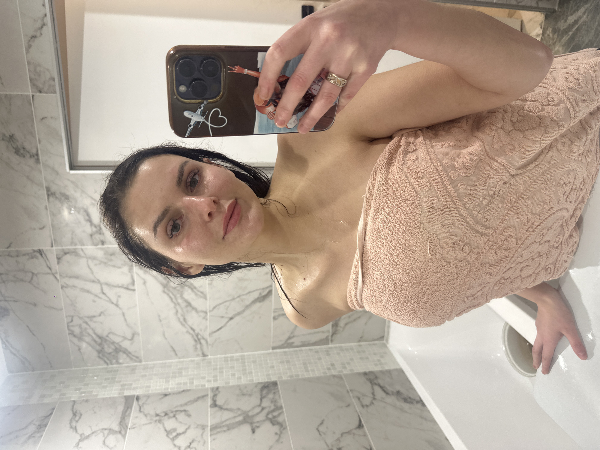 A person is taking a selfie in a bathroom. The person has dark hair and is wearing a pink towel. Their face is partially visible, and they are holding a smartphone with a phone case that has a design on it. The background of the image shows marble tiles on the walls and a white bathtub. The lighting suggests that it could be daytime or well-lit artificial lighting. The person's expression is neutral, and the overall scene appears to be a casual moment captured in a private setting. There is no visible text in the image.
