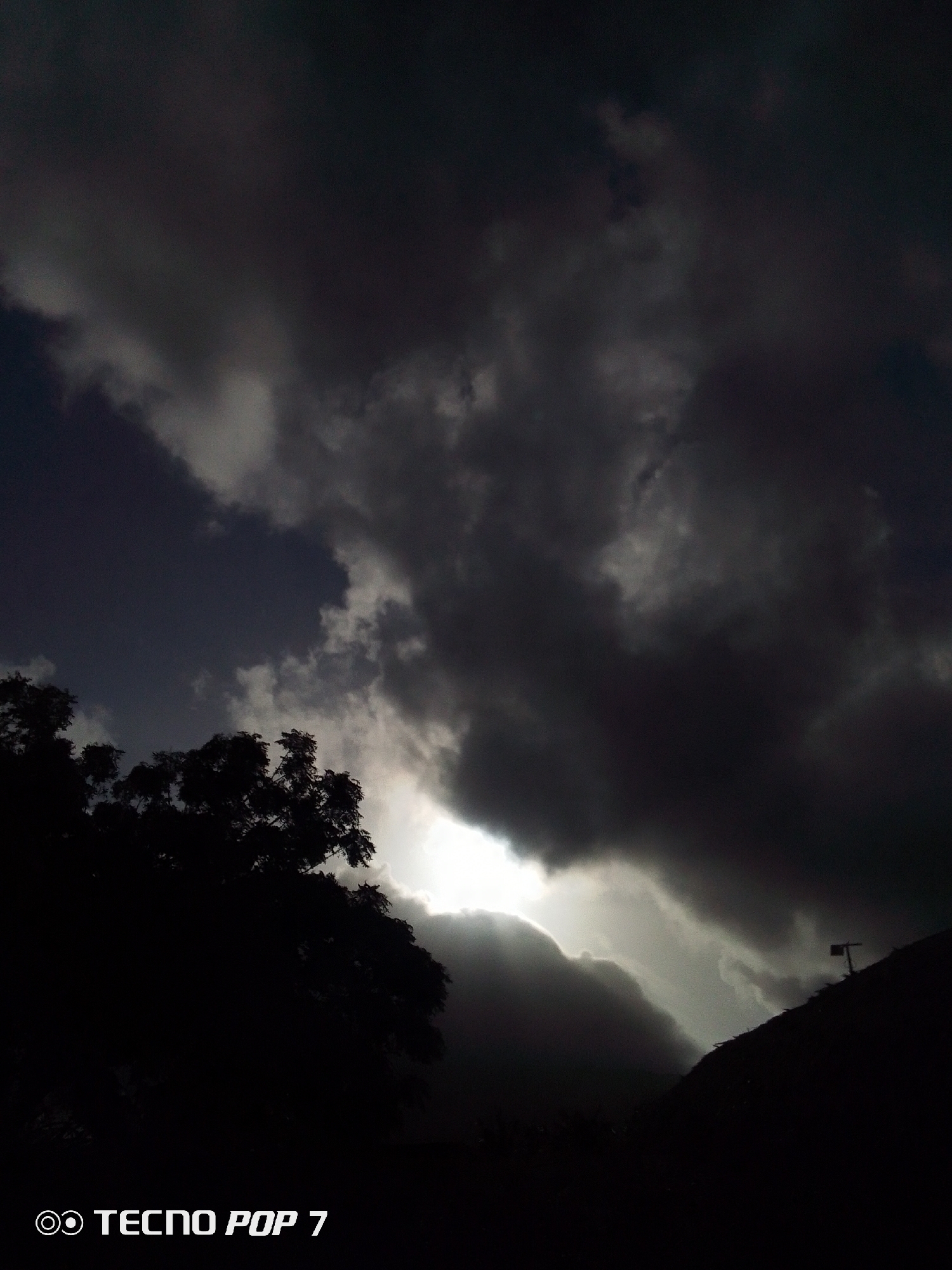 The image depicts a dramatically dark sky filled with heavy, ominous clouds. A bright opening in the cloud cover reveals sunlight, suggesting the potential for a break in the storm. In the lower left of the frame, the silhouette of a tree is visible against the stormy sky. In the lower right, there is a dark silhouetted hill or building with what appears to be a small structure on top. The photograph was taken with a TECNO POP 7 mobile phone, presumably in Pedro González, Venezuela.