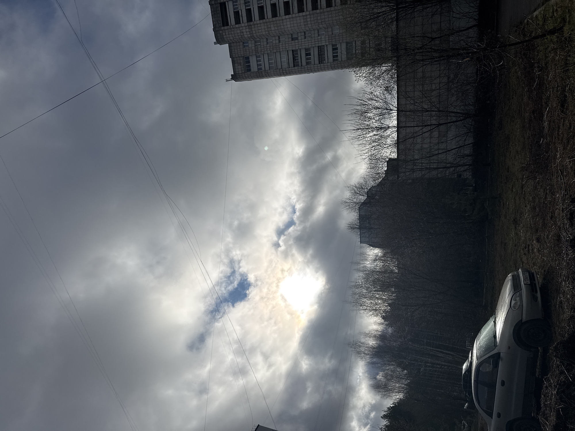 The image shows a cloudy sky in the city of Chaikovsky, Russia. The sun is obscured by thick clouds, with only a bright spot visible. Power lines crisscross the sky. A tall, grey, Soviet-era apartment building is visible in the upper portion of the image. Below, bare trees line the horizon. A silver car is parked on a patch of brown grass and dry vegetation. The overall impression is one of urban decay and a somber, overcast day.