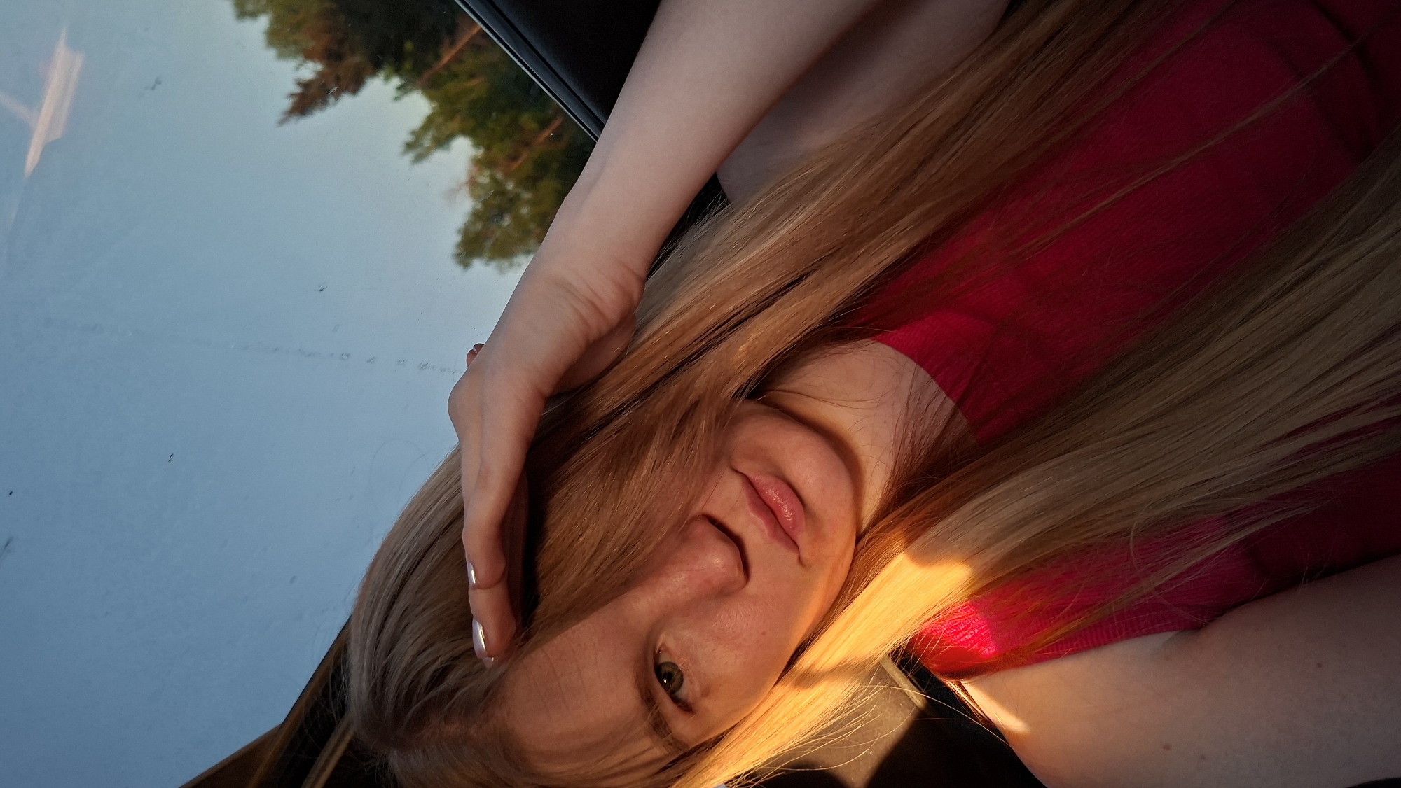 A woman with long blonde hair, wearing a red shirt, is pictured from a low angle. She has one hand on her head, and her face is partially obscured by her hair. The background is a sky and some trees, reflected in what appears to be a car window. The location is Chaikovsky, Russia, based on external information.