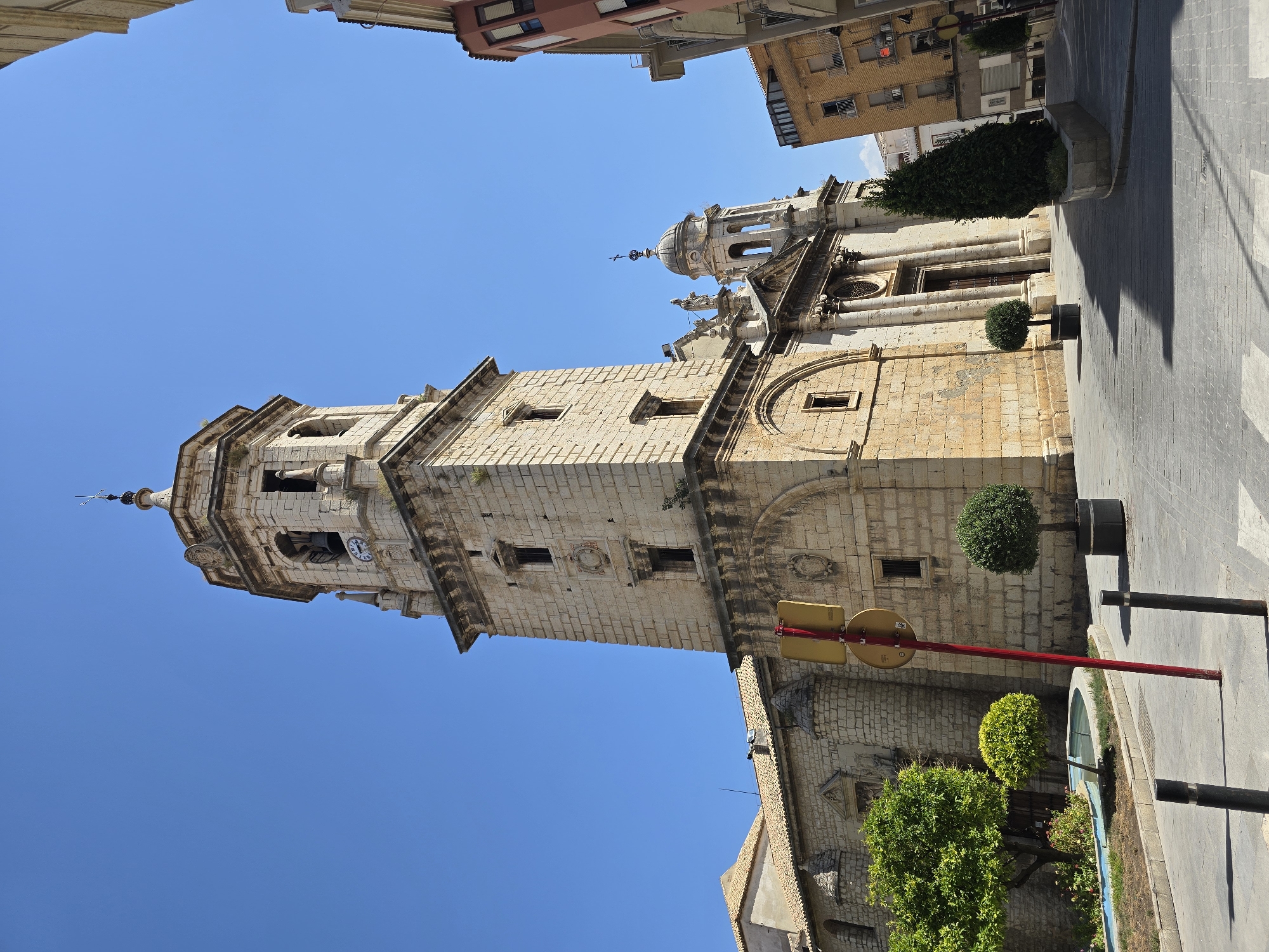 This image depicts the exterior of a historic stone church, likely a cathedral or significant religious building, in Jaén, Spain. The church features a tall, multi-tiered bell tower with a clock face visible near the top. Adjacent to the tower is the main facade of the church, characterized by classical columns and an ornate entrance. The building is constructed of light-colored stone blocks.
In the foreground, a paved plaza area is visible, with white pedestrian crossings painted on the ground. Several potted trees and a small, curved fountain with blue water are placed around the plaza, adding greenery to the urban setting. A red traffic signpost with a yellow circular sign is prominently located in the mid-ground, leaning slightly.
Surrounding the church and plaza are other buildings, including a multi-story residential or commercial building with balconies on the right side of the frame. The sky is clear and bright blue, suggesting a sunny day. The architectural style and the presence of Spanish signage (though not clearly legible in detail) strongly indicate a location within Spain, and specifically, the style aligns with many historical churches found in Andalusian cities like Jaén. The scene appears to be a public square adjacent to a religious site, likely a central area of a town or city. There are no people visible in the image.