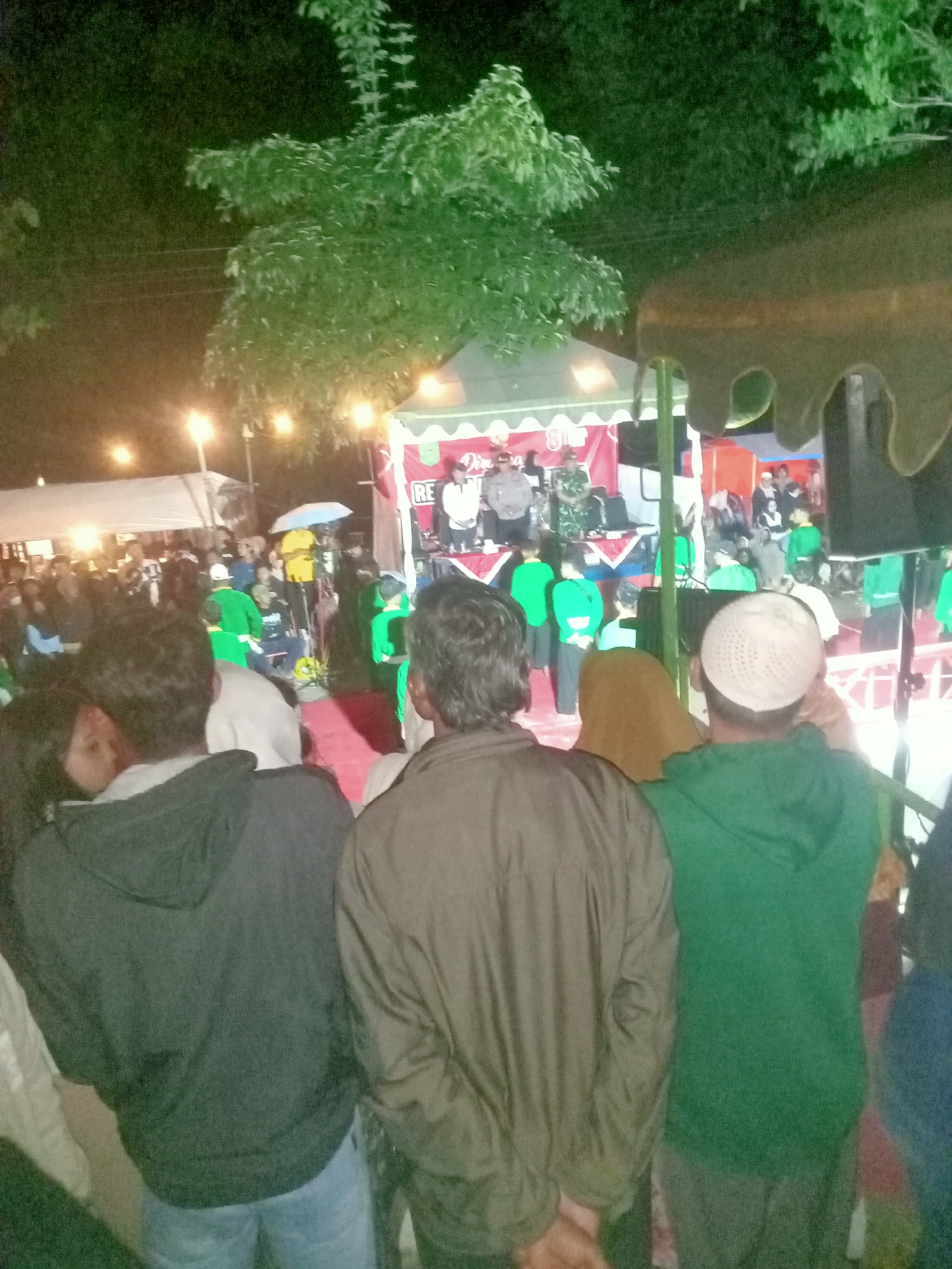 In Pule, Indonesia, an outdoor event takes place at night. A stage is set up with people on it, including individuals in military uniforms and others in formal attire. A red banner with white lettering is visible behind them. A crowd of people stands in front of the stage, some holding umbrellas. The foreground features the backs of three individuals, two wearing hoodies and one a jacket. The environment is dimly lit, with string lights illuminating the scene. Trees are visible in the background.