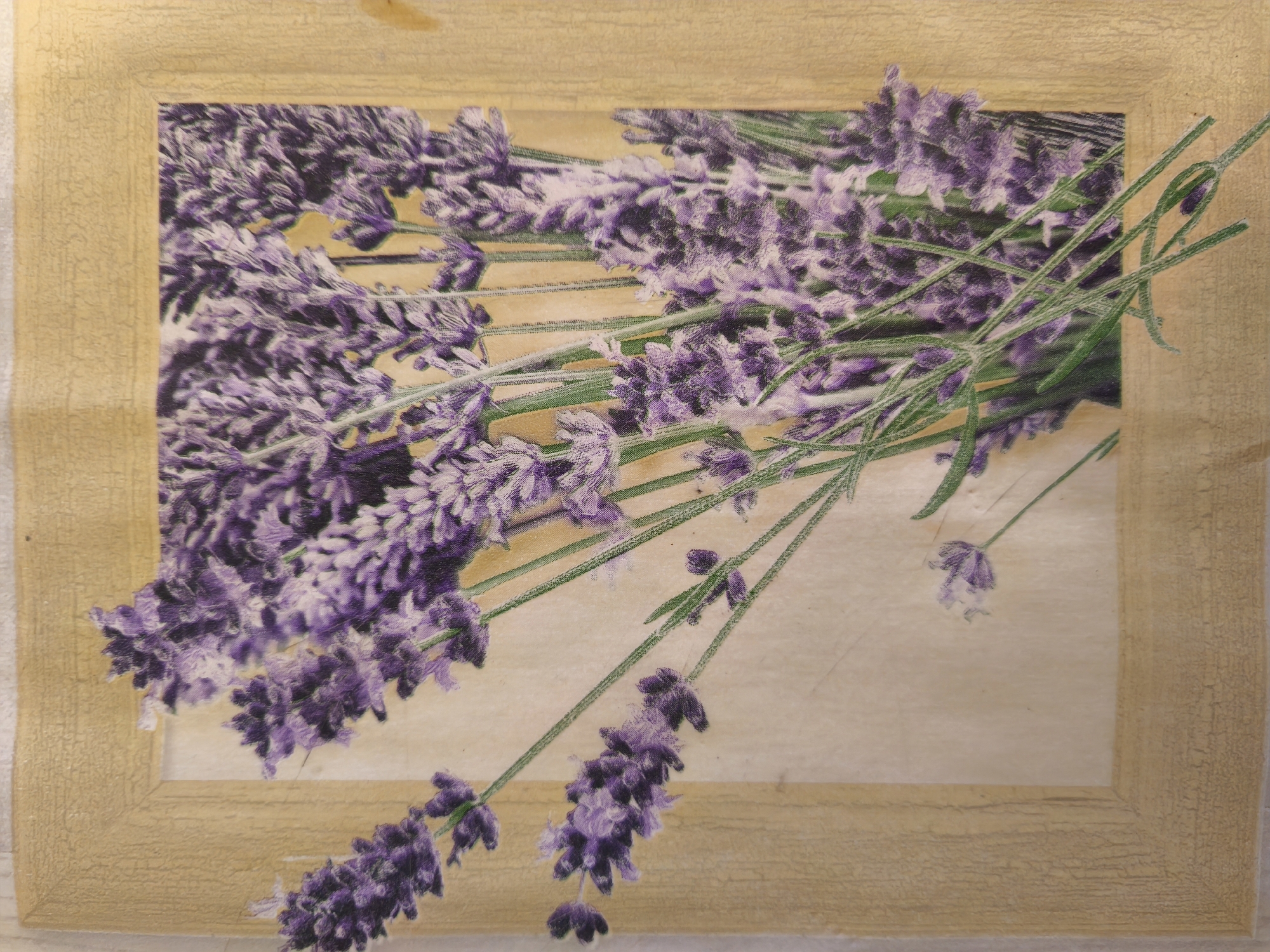 The image shows a close-up, overhead view of a framed picture featuring a bouquet of lavender flowers. The frame is made of a textured, light-brown material with a distressed finish, and it encloses a light-colored background, possibly paper or canvas, upon which the lavender is depicted. The lavender stems are a vibrant green, and the flowers themselves are a rich purple, arranged in a cascading fashion. The artwork appears to be a print or decoupage applied to a flat surface. There are no people visible in the image, and no specific location in Leninsk-Kuznetsky, Russia can be identified from the artwork itself.