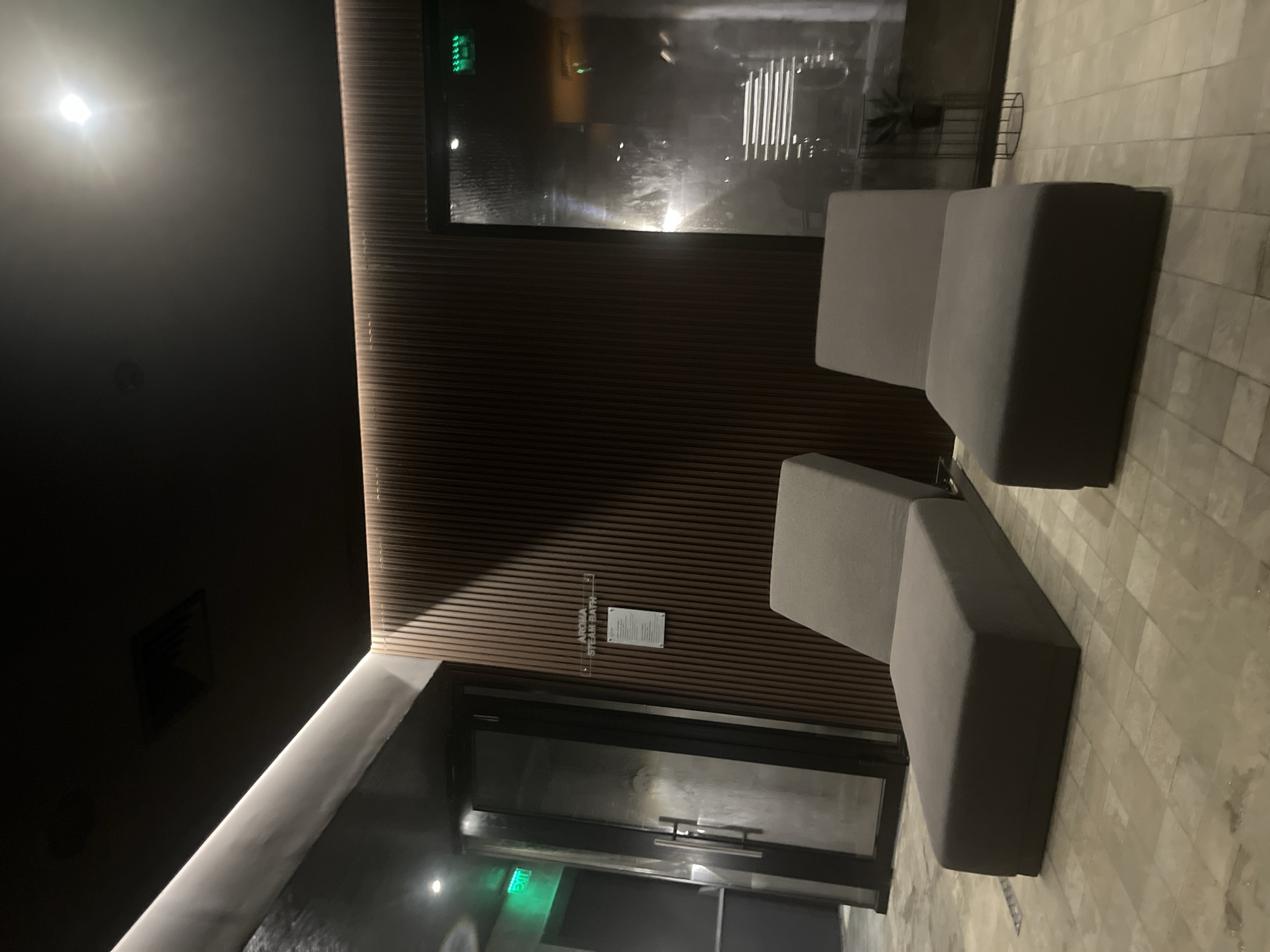 The image depicts an interior space, potentially a spa or wellness center, in an unknown location but purportedly in Sofia, Bulgaria. The scene includes modern, minimalist design elements. There is a dark ceiling with a recessed light. One wall is clad in a horizontally-striped wood-like material.
There are two gray modular seating units arranged in a staggered fashion, suggesting a waiting area. Behind the wall is what appears to be a door or entrance to a "Steam Bath" area, indicated by signage near the door. There's also a window behind the seating, reflecting light. The floor is tiled and appears to be stone.