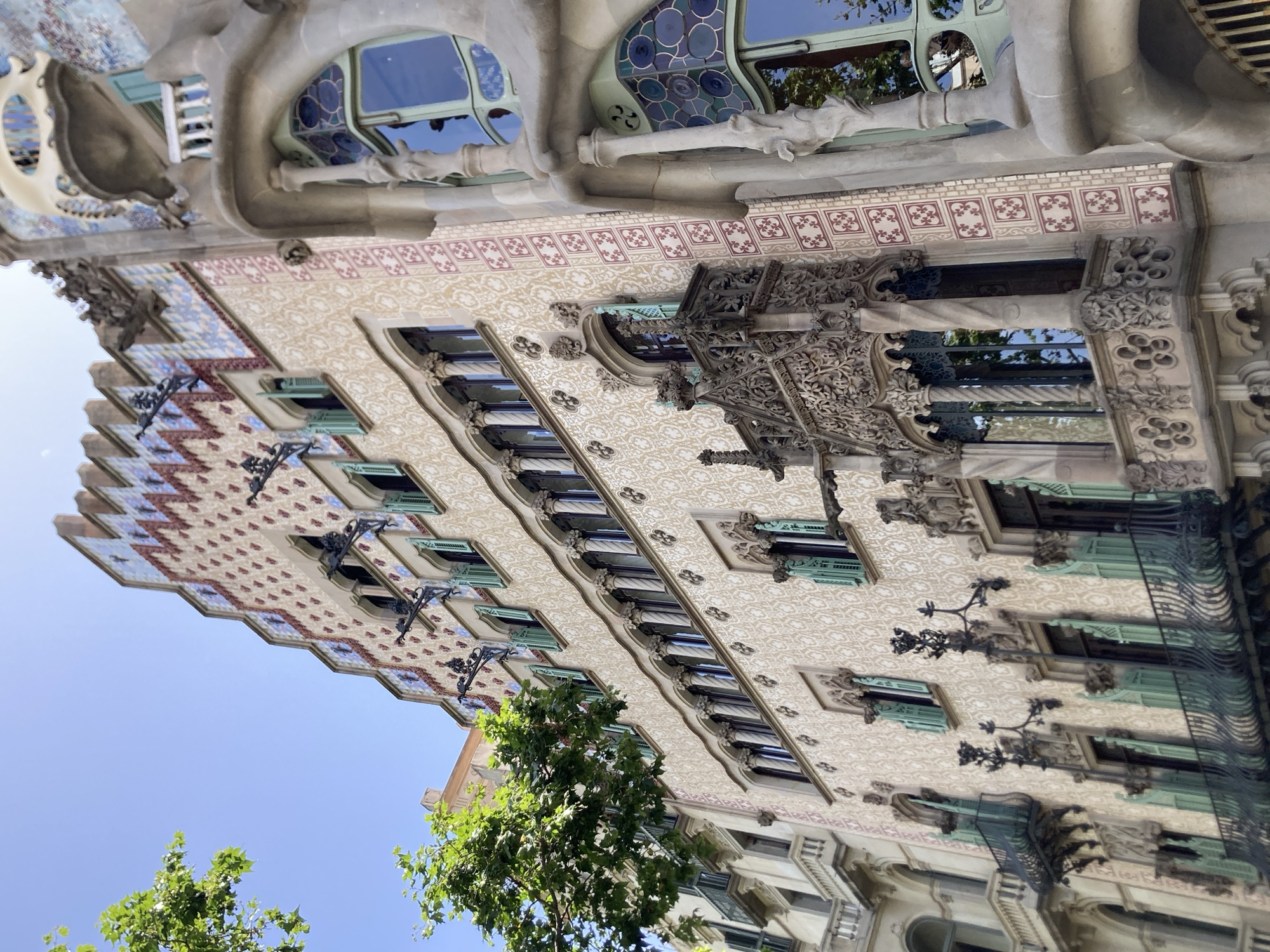 This is a low-angle, close-up shot of the facade of a building with ornate architectural details, bathed in bright daylight under a clear blue sky. The building's exterior is a mosaic of intricate patterns and textures, featuring a repeating geometric design in various colors, including light beige, pink, and blue tiles. Several windows with light teal shutters are visible, some partially open. Decorative elements, such as sculpted gargoyles and wrought-iron balconies with delicate scrollwork, are integrated into the facade. The building's structure itself has organic, flowing lines, reminiscent of natural forms, with curved surfaces and elaborate stonework.
In the foreground, lush green trees with vibrant foliage are partially visible, their leaves catching the sunlight. The overall impression is one of artistic craftsmanship and historical significance, suggesting a location known for its unique architecture. Given the location context of Barcelona, Spain, this building is likely a landmark of Catalan Modernism, possibly designed by Antoni Gaudí, given the characteristic organic shapes and decorative tilework. The scene is peaceful and serene, with no people or activity explicitly depicted, focusing solely on the architectural splendor and the interplay of light and shadow on the building's surface. The time of day appears to be midday or early afternoon due to the strong sunlight and clear sky, indicating fair weather.
