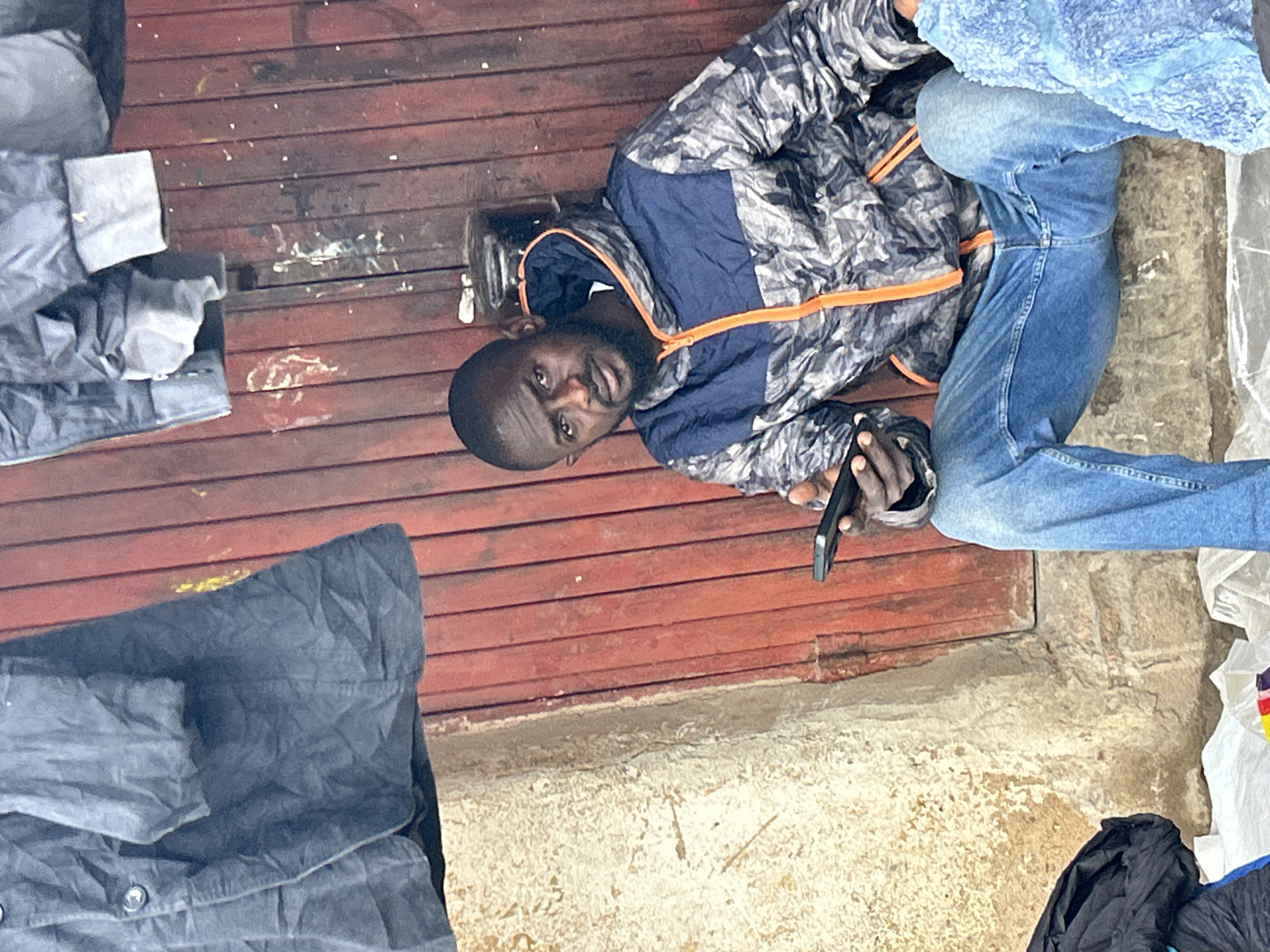 A Black man with a beard and dark skin is sitting on a concrete ledge in front of a red, corrugated metal door. He is wearing a camouflage patterned jacket with blue and orange accents, blue jeans, and a grey hooded shirt underneath the jacket. He is holding a black smartphone in his right hand and looking towards the camera with a neutral expression. The man appears to be outdoors, with a textured concrete wall to his left and various items, including a black bag and white plastic sheeting, on the ground in front of him. The background includes what appears to be hanging garments or fabric, possibly coats, made of dark, possibly denim or heavy cotton material. The scene is likely located in a market or outdoor stall area in Jos, Nigeria.