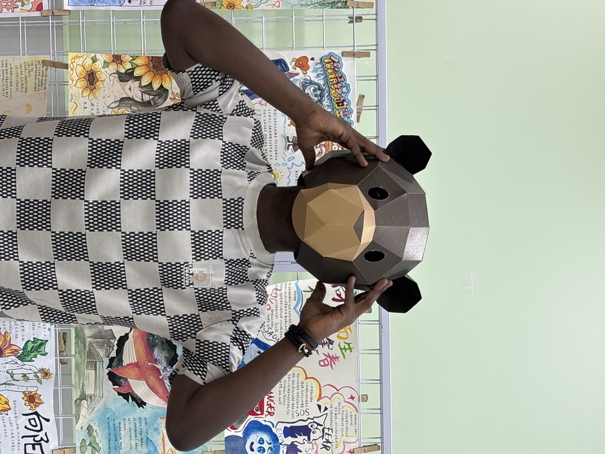 The image, captured in Jiangjin, China, shows a person holding a three-dimensional, polygonal papercraft bear mask. The mask is predominantly dark brown with a lighter brown snout and black ears, covering the person's entire face. The individual wears a short-sleeved shirt with a black and white checkered pattern, and a black beaded bracelet is visible on their right wrist.
The background features a light pastel green wall, against which a metal grid display is set up. Numerous artworks, appearing to be drawings or paintings, are attached to this grid with wooden clothespins. These artworks are colorful and varied, depicting scenes like sunflowers, a whale, and abstract designs. Many of the pieces include prominent text in Chinese characters. English words such as "ANGER," "FEAR," "ENYY" (likely "ENVY"), "SOS," and "SADA" are also visible on some of the artworks, often integrated into emotional or thematic illustrations. One piece explicitly mentions "心理健康的重要性" (The importance of mental health). The setting suggests an indoor educational or exhibition space, possibly displaying student artwork. The image itself is rotated 90 degrees clockwise.