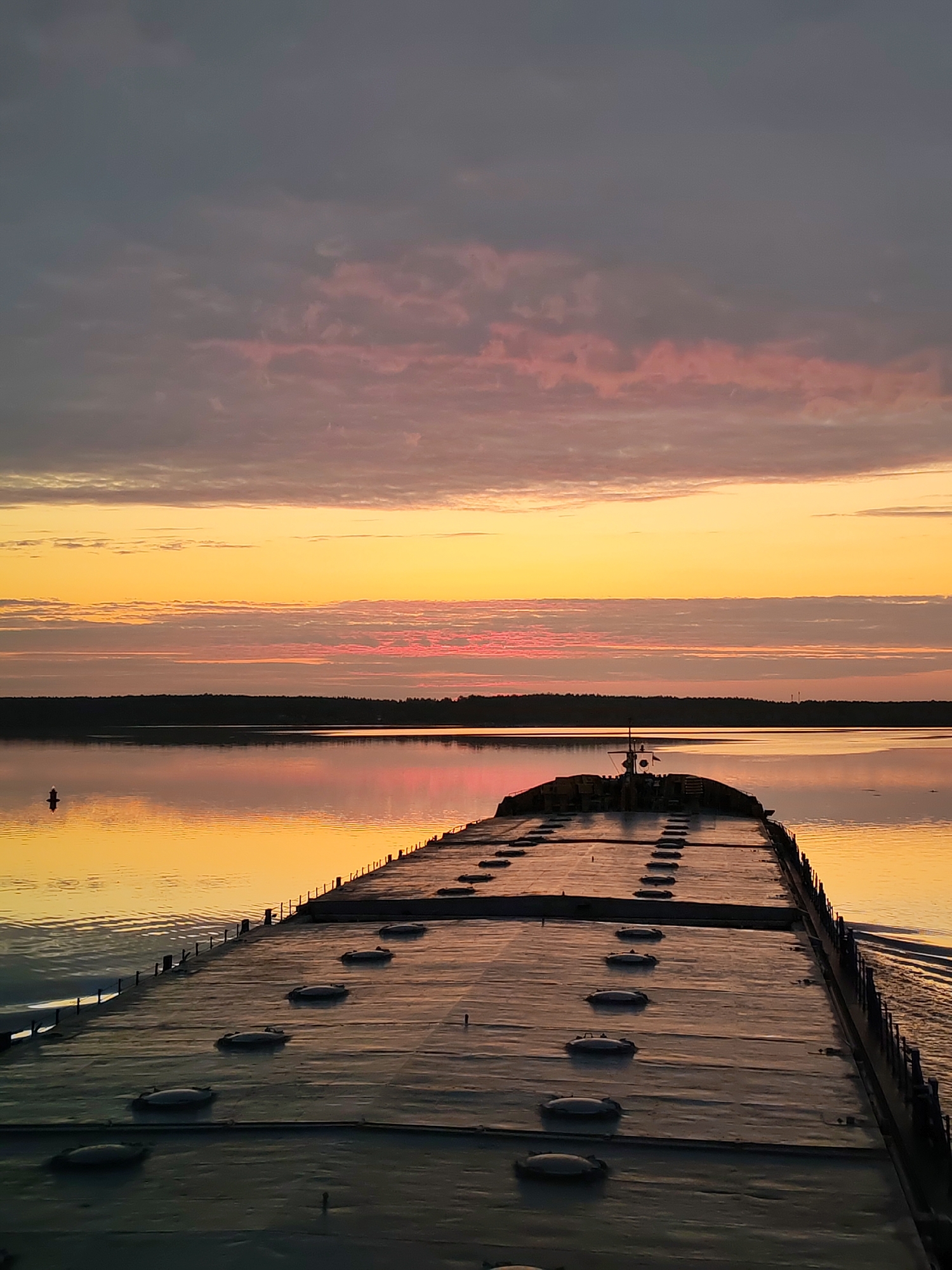 A long, flat barge sits on the water at sunset. The barge fills most of the foreground. A row of round hatches runs down its length. The water is calm and reflects the golden light of the setting sun. The sky has a mix of colors, including dark gray, pink, and bright yellow. A line of trees appears on the horizon, and a buoy bobs in the water to the left. The location is in the city of Unknown, Unknown.