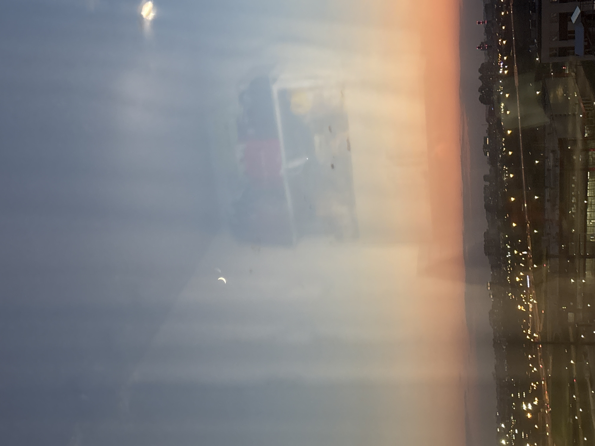 The image depicts a partial solar eclipse viewed through a window. The sky is overcast, with a visible crescent shape where the sun is obscured. Reflected in the window is a blurred cityscape at night with numerous lights, possibly the city of Murino, Russia. The city lights form a backdrop to the eclipse visible above.