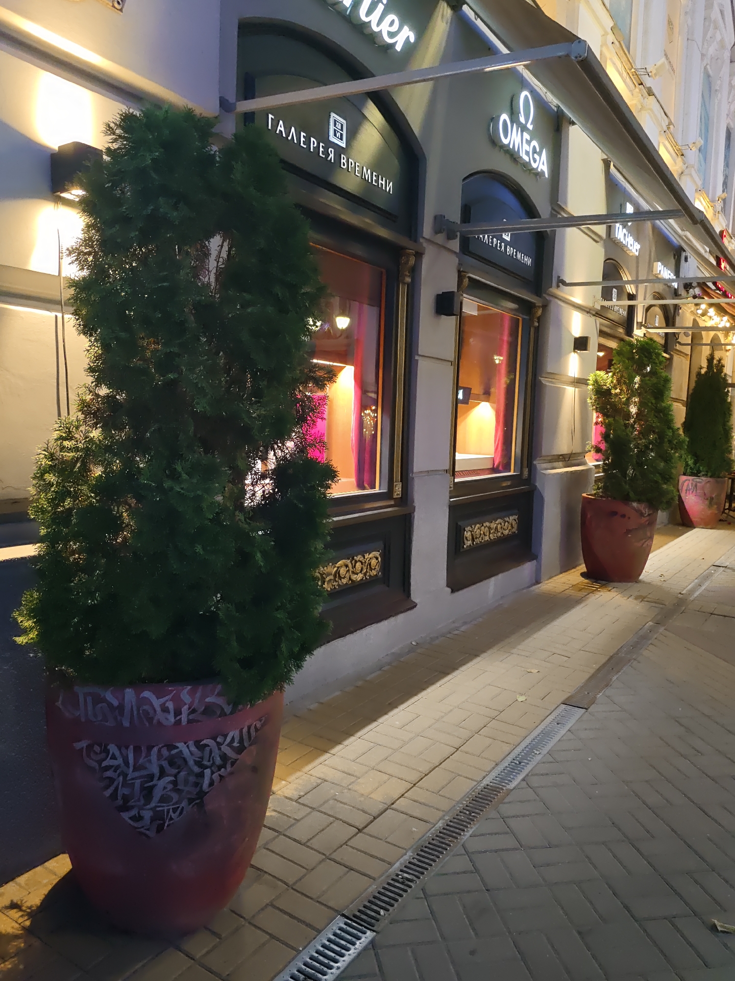 The image depicts the exterior of a luxury watch boutique, likely "Omega" or a similar high-end establishment, located in Rostov-on-Don, Russia. A large evergreen tree in a decorative red planter is prominent in the foreground, with visible graffiti-like markings on the planter. Behind the tree, the storefront features large display windows with plush purple curtains visible inside. Above the windows, a sign reads "ГАЛЕРЕЯ ВРЕМЕНИ" (Gallery of Time) and displays the Omega logo, confirming it as an Omega boutique. The building's facade is well-lit by wall-mounted sconces, casting dramatic shadows. The street is paved with interlocking bricks, and a drainage grate runs along the sidewalk. Another similar evergreen tree in a red planter is visible further down the street, suggesting a consistent design choice for the storefront's landscaping. The overall impression is one of a well-maintained, upscale commercial establishment at night.