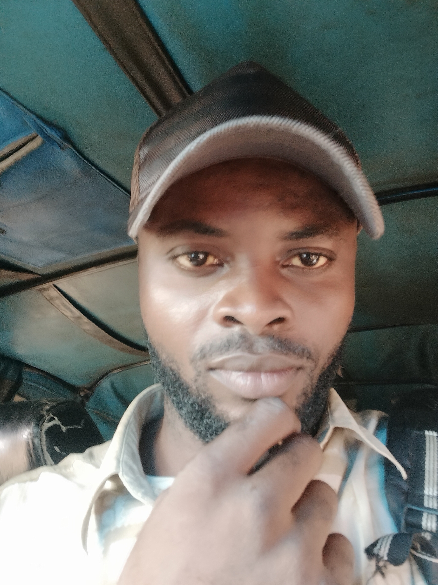 A man is sitting inside what appears to be a "Keke Napep" or a tricycle taxi, a common mode of transportation in many parts of Nigeria. The man is wearing a baseball cap. He has dark skin, short dark hair and beard, and is wearing a collared shirt. He is touching his chin with his hand. The interior of the tricycle is visible, with the ceiling appearing to be lined with fabric. The photograph was likely taken in Ikorodu, Nigeria, as this type of transportation is common in that area.