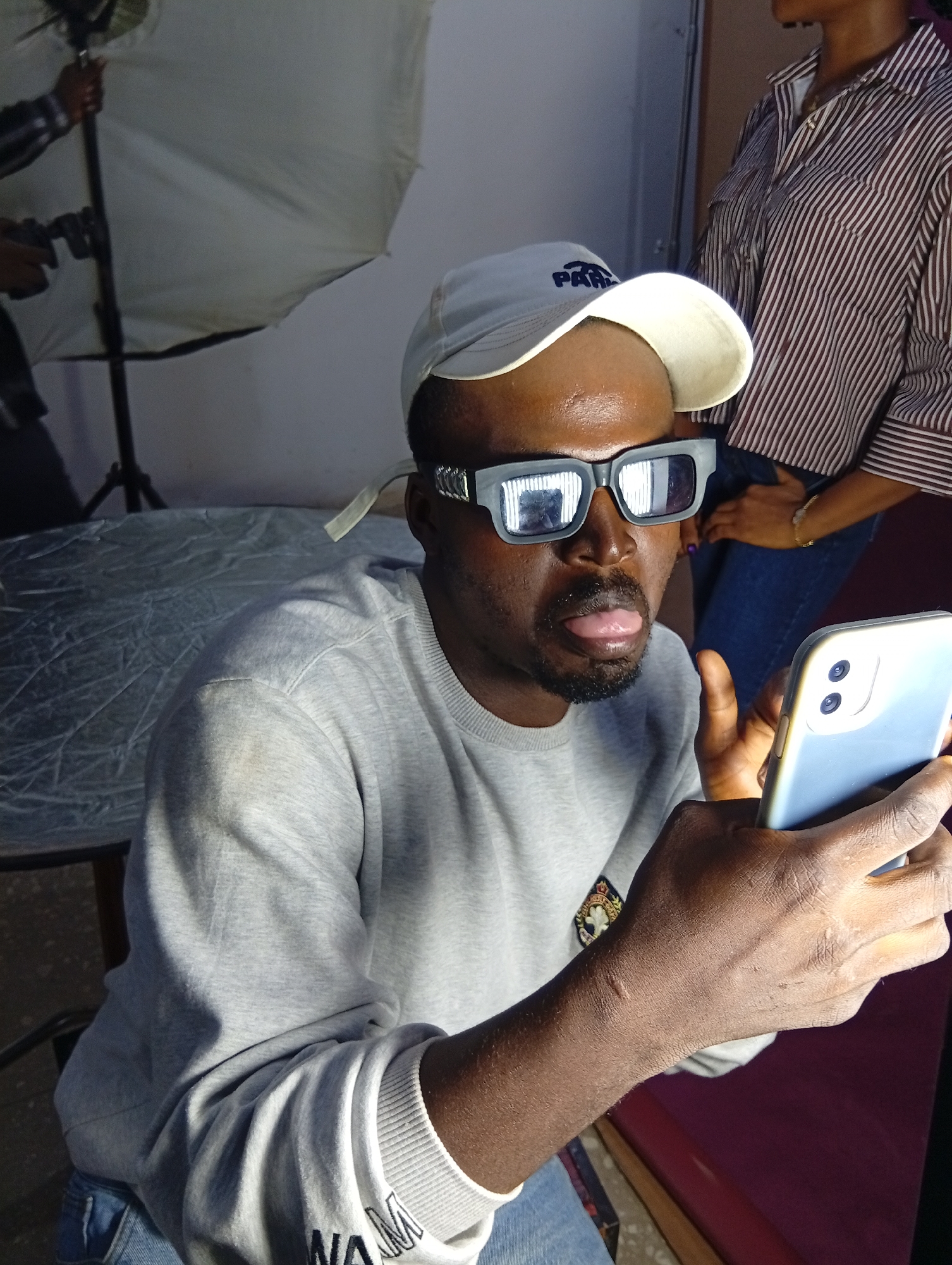 The image shows a medium shot of a Black man wearing a light-colored baseball cap, gray sweatshirt, and blue jeans. He is also wearing distinctive rectangular sunglasses with reflective lenses. The man is holding a light blue smartphone in his right hand and appears to be looking at it, with his tongue slightly sticking out. The camera angle is from the man's left, at chest level.
In the background, a person wearing a striped shirt is visible, along with photographic equipment, including a light stand and a large diffuser. This suggests the scene is likely a studio or a prepared photoshoot setting. The lighting appears to be artificial, casting shadows and highlighting the subjects. The overall mood is casual and perhaps playful, given the man's expression and the context of a photo shoot. There is no clear indication of the time of day or weather, and the location is unknown.
Visible text includes "PARIS" on the baseball cap. The sweatshirt has a circular emblem on the left chest. The phone case is plain light blue.