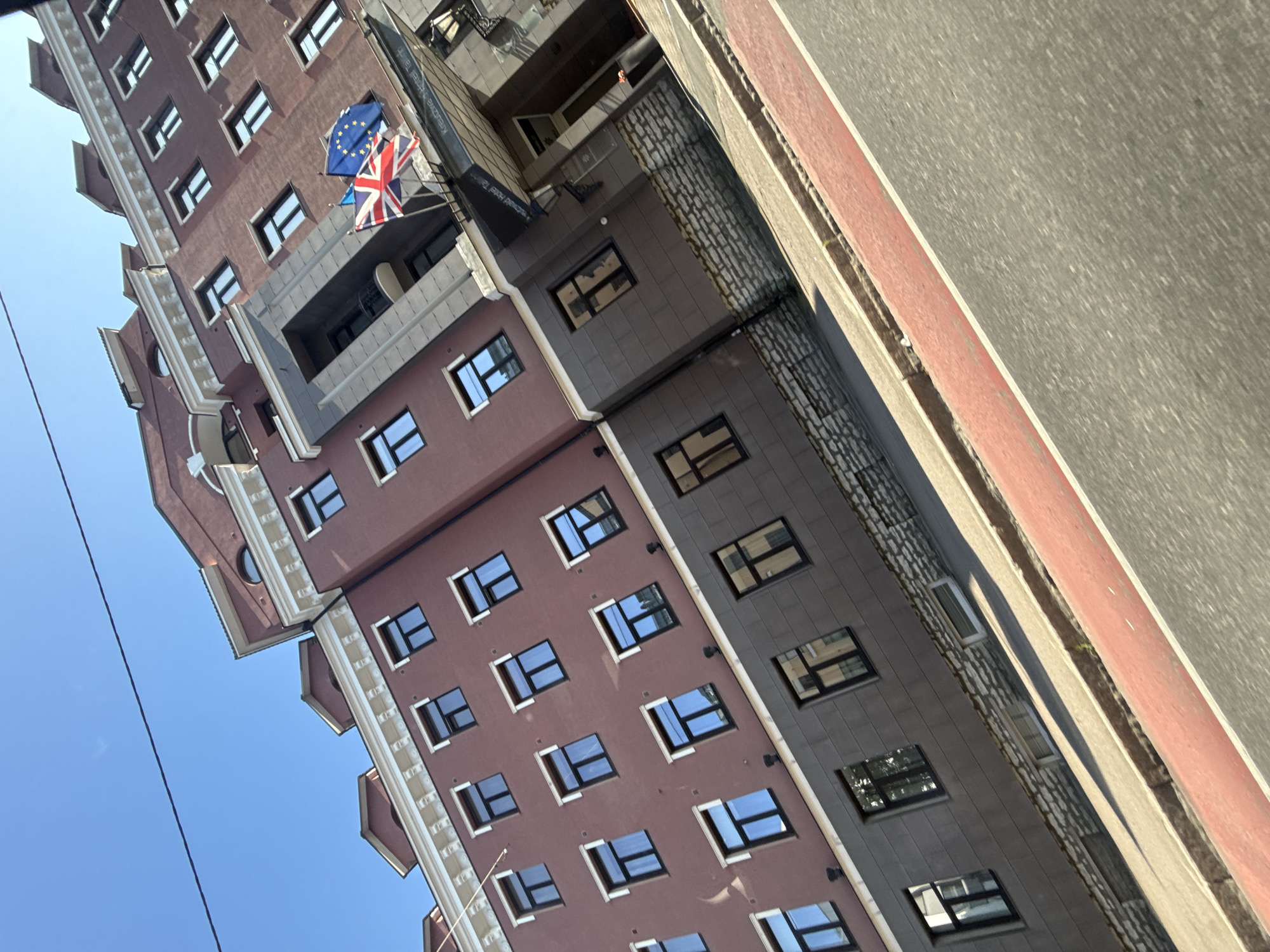 The image shows a multi-story building with a red brick facade and dark gray lower levels, tilted at an angle. The building has numerous rectangular windows with dark frames, arranged in a grid pattern. At the entrance, there is a canopy with the text "Kreutzwald Hotel Tallinn" visible, flanked by the Union Jack and the European Union flag. The building is situated in Tallinn, Estonia. In the foreground, a sidewalk with a red-striped lane runs alongside the road, which is paved and appears to be asphalt. The sky is clear and blue, with a thin black wire visible across the top of the frame. No people are clearly visible in the image.