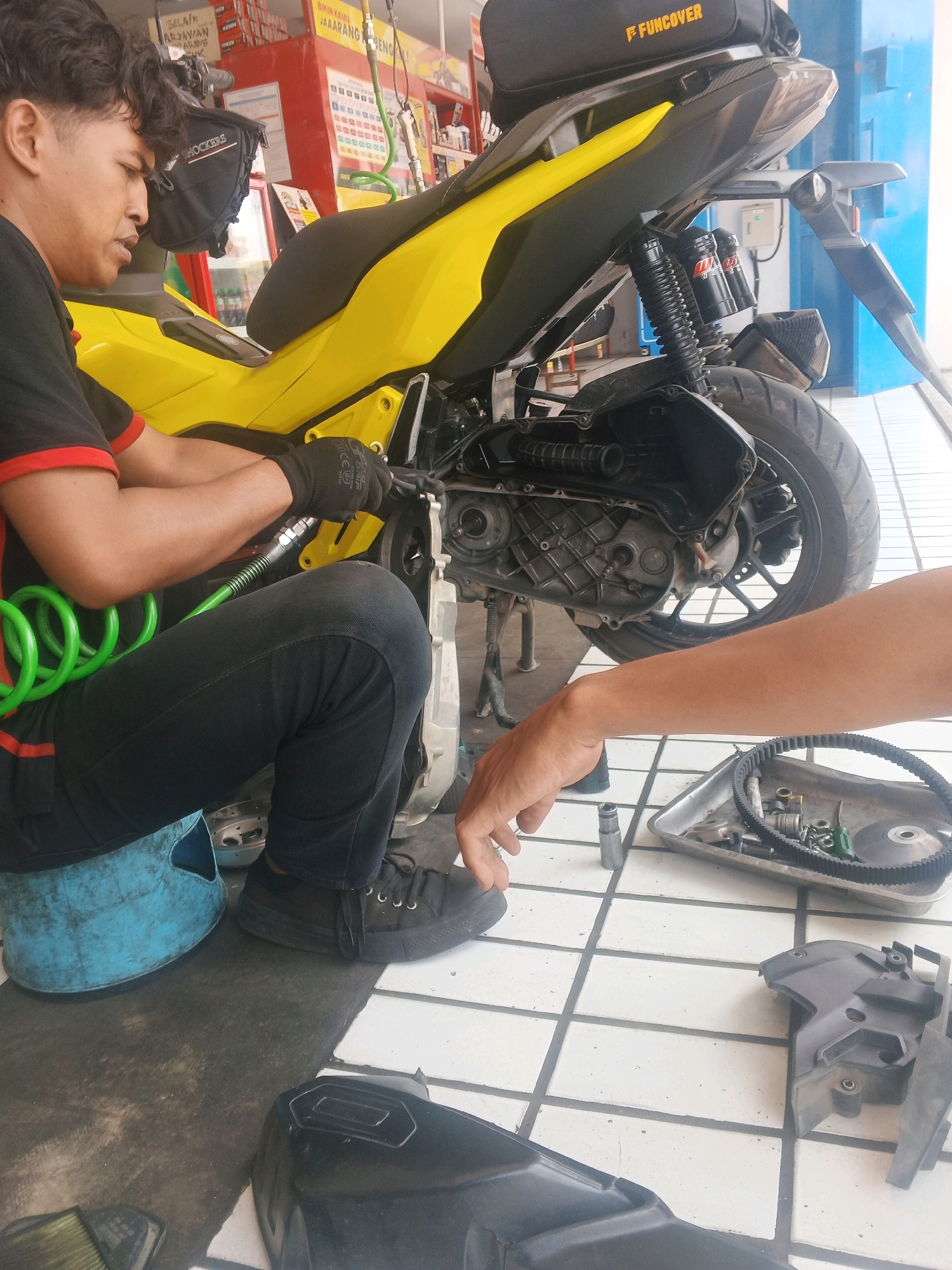 A person wearing black gloves and black pants is working on the engine of a yellow and black motorcycle in an outdoor garage or workshop. The person is seated on a blue plastic stool and appears to be using tools to repair the motorcycle. There are various motorcycle parts and tools scattered around the scene, including a metal tray with a belt and other small components, and a black plastic fairing piece on the ground. The floor is tiled. In the background, there are shelves with products and signage, suggesting a commercial establishment. The overall setting appears to be a repair shop in the city of Taman, Indonesia.