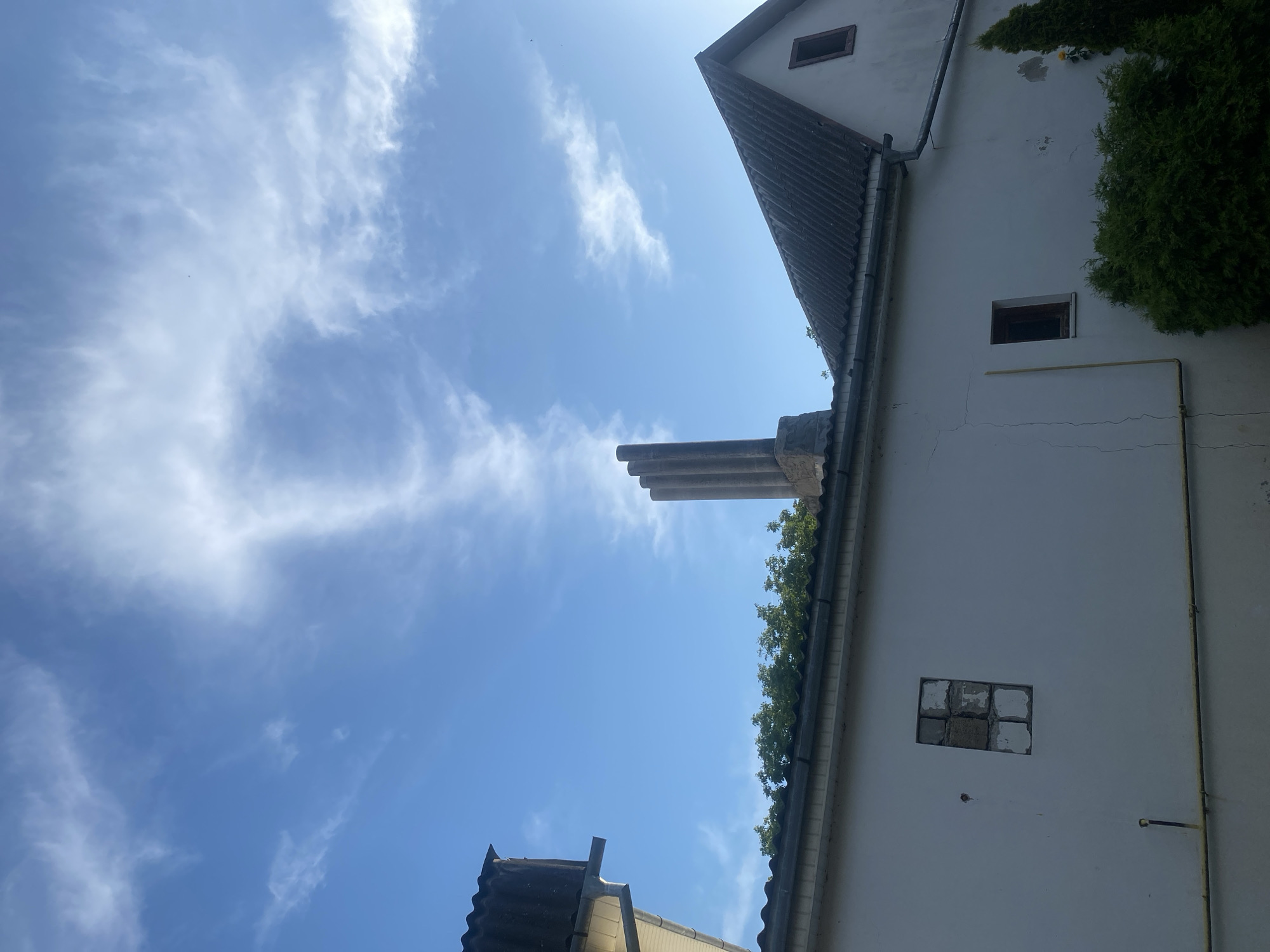 The image captures a close-up view of a building's exterior against a bright blue sky with scattered white clouds. The building features a white, stucco-like wall with some visible cracks. There are two windows on the wall; one is a small, broken window with exposed brickwork, and the other is a smaller, intact window with dark wood trim. A yellow pipe runs horizontally across the wall.
The roof of the building is visible, covered with dark, corrugated material. A prominent chimney with three cylindrical pipes stands on the roofline. Adjacent to the chimney is a section of dense green foliage, likely trees or bushes. Another building section with a gray, sloped roof and a downspout is partially visible on the left side of the image. The overall scene suggests an outdoor view of a residential or industrial structure, possibly in a suburban or rural setting.