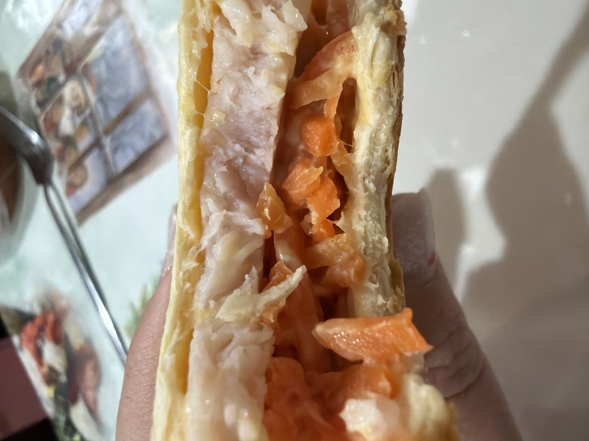 The image shows a close-up of a pastry, possibly a turnover or pie, being held by a hand. The pastry has a golden-brown crust and is filled with a mixture of shredded light-colored meat (likely chicken or turkey) and orange-colored shredded vegetables (possibly carrots). The setting appears to be indoors, with a table visible in the background, including a plate with a blurred image of food packaging and a spoon. No further information available to determine the location in the city of Chaikovsky, Russia.
