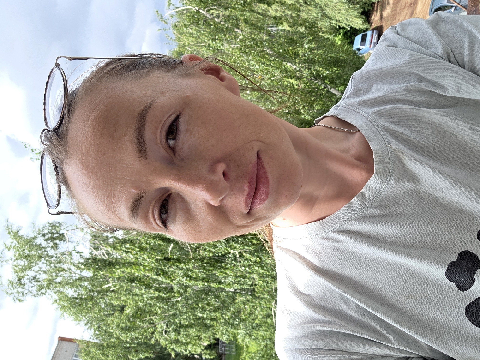 A medium-shot portrait, taken from an elevated position, shows a woman with fair skin and freckles looking directly at the camera with a slight smile. Her hair is blonde, pulled back, with glasses resting on her head. She wears a light-colored t-shirt and a thin silver necklace is visible at her neck. Behind her, a vibrant green leafy tree fills the background. The sky is visible above the tree, suggesting an outdoor setting. In the distance, elements of a cityscape can be seen, including a building and a blue car. The specific location within the city is Unknown, Unknown.