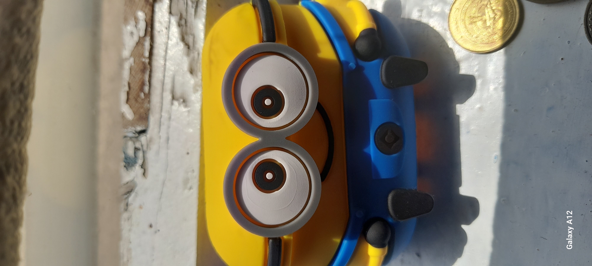 In a close-up, eye-level shot, a yellow Minion toy is positioned on a weathered, white ledge. The toy, which appears to be a container with a lid, features large, round goggles with white irises and black pupils, and a blue fabric-like garment. The toy's feet are black and rounded, and on its blue garment, there's a circular emblem with a symbol resembling a stylized "G". To the right of the toy, two coins are partially visible. The background is out of focus, but suggests a rough, textured surface. The image appears to have been taken outdoors, possibly in a location with natural lighting. The "Galaxy A12" watermark is present in the bottom left corner. This image is not located in Tash-Dobo, Kyrgyzstan.