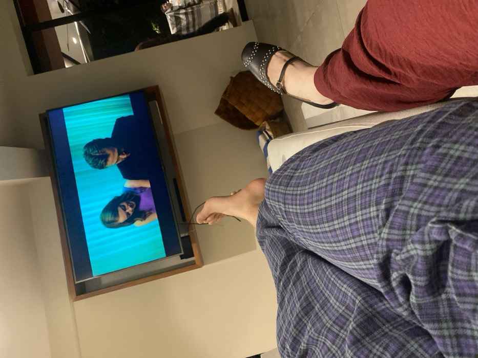 A low-angle shot depicts two people relaxing, likely indoors. One person wears burgundy pants and black, studded sandals. The other is in blue and purple plaid pajamas. They appear to be sitting on a white couch. A television mounted on the wall shows a scene with a man and a woman. Glasses lay next to the leg of the person wearing the pajamas. A brown cushion is visible on the couch behind the person in burgundy.
