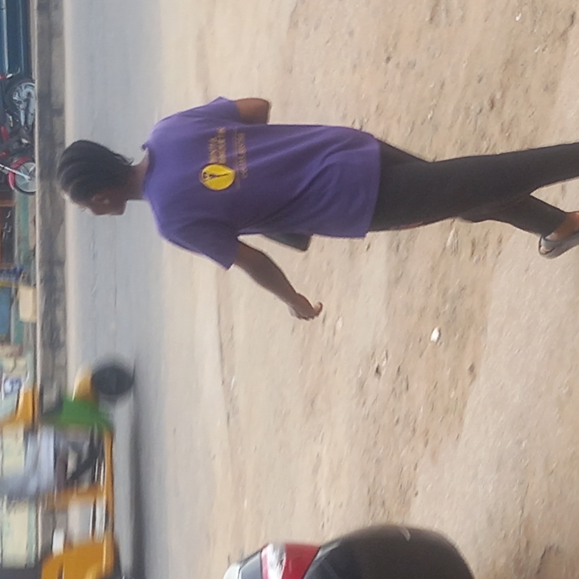 A person in a purple t-shirt and dark pants walks along a dusty, likely unpaved, roadside. The person is wearing sandals. Yellow auto rickshaws and motorcycles are visible near the side of the road. The location is Dadi Riba, Nigeria.