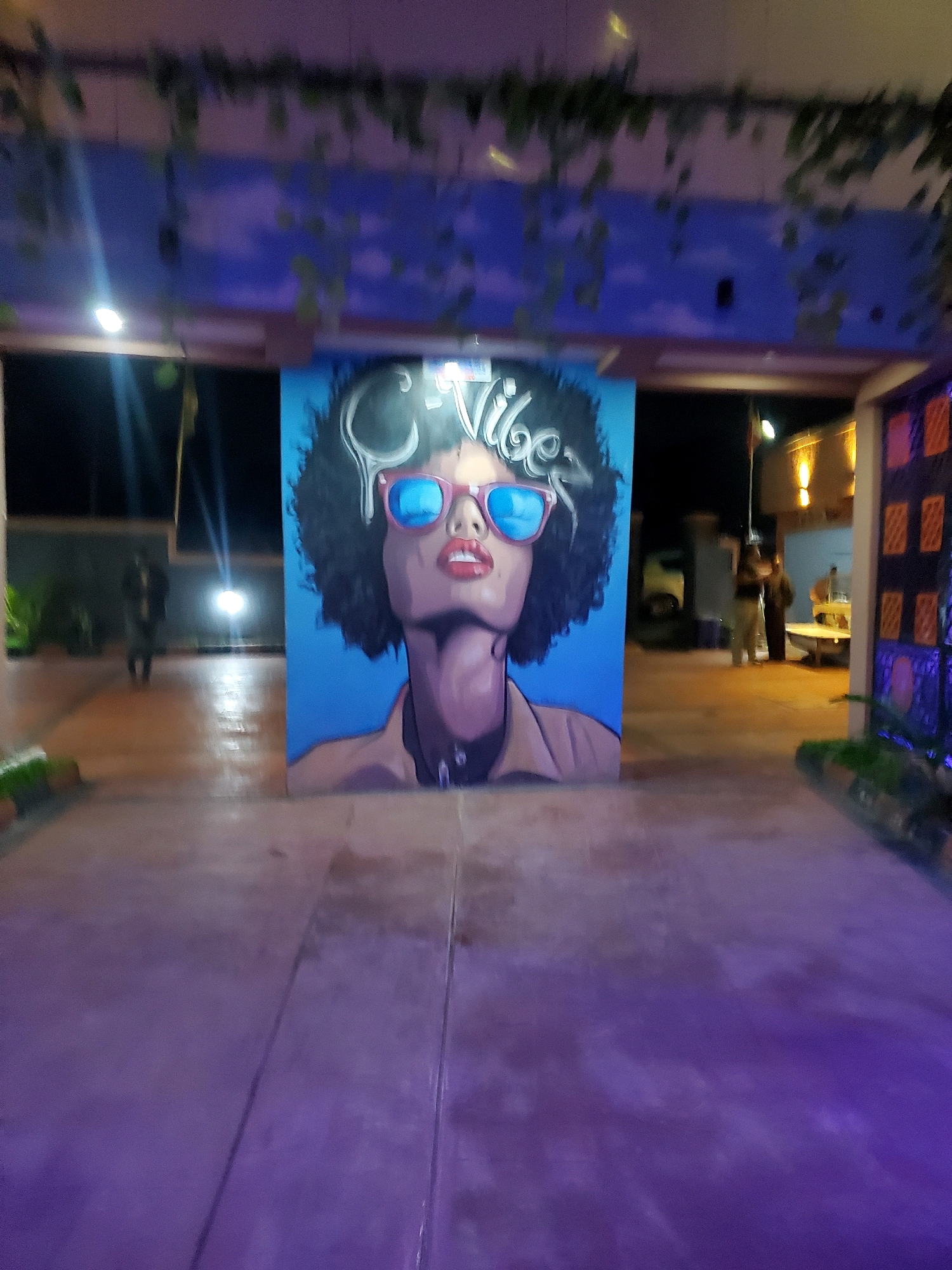 A mural of a woman with an afro and sunglasses is displayed on a blue background, with the word "Vibes" written above her head. The mural is placed in a pathway area with purple lighting. In the background, several people are visible walking and standing, and a yellow vehicle is parked. The scene appears to be outdoors at night. It is not possible to determine the specific location in Jos, Nigeria from the image.