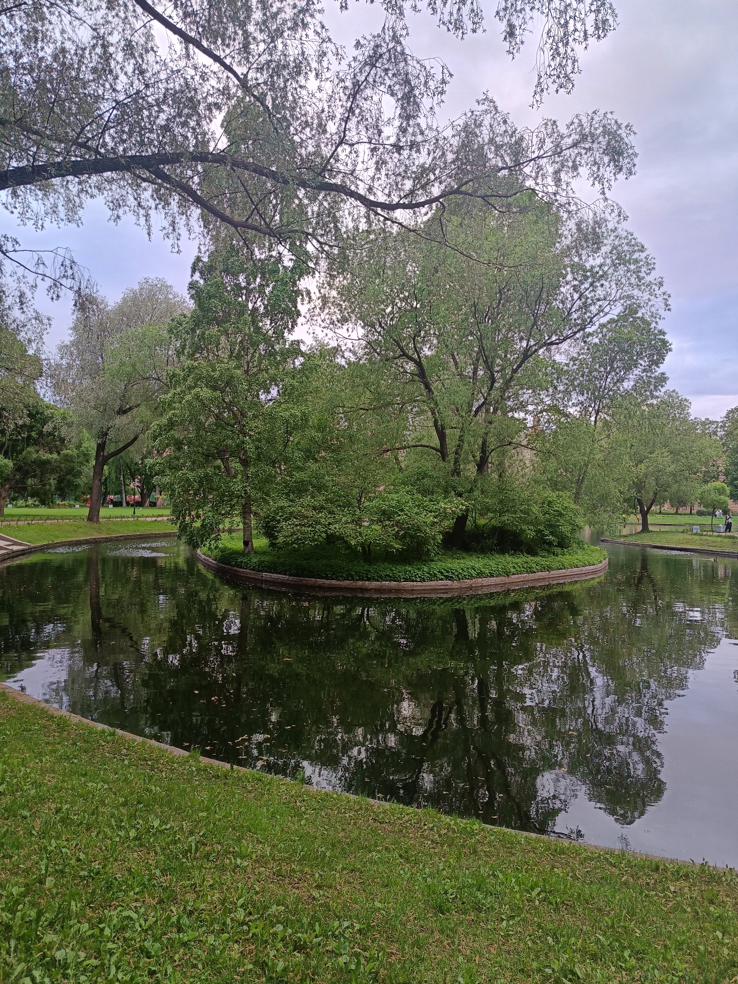 The image shows a circular island with trees and bushes in the middle of a pond. The pond reflects the trees and the sky. Green grass surrounds the pond. Branches of a tree hang over the pond in the foreground. In the background, there are more trees and some people walking in the park. The location is Saint Petersburg, Russia.