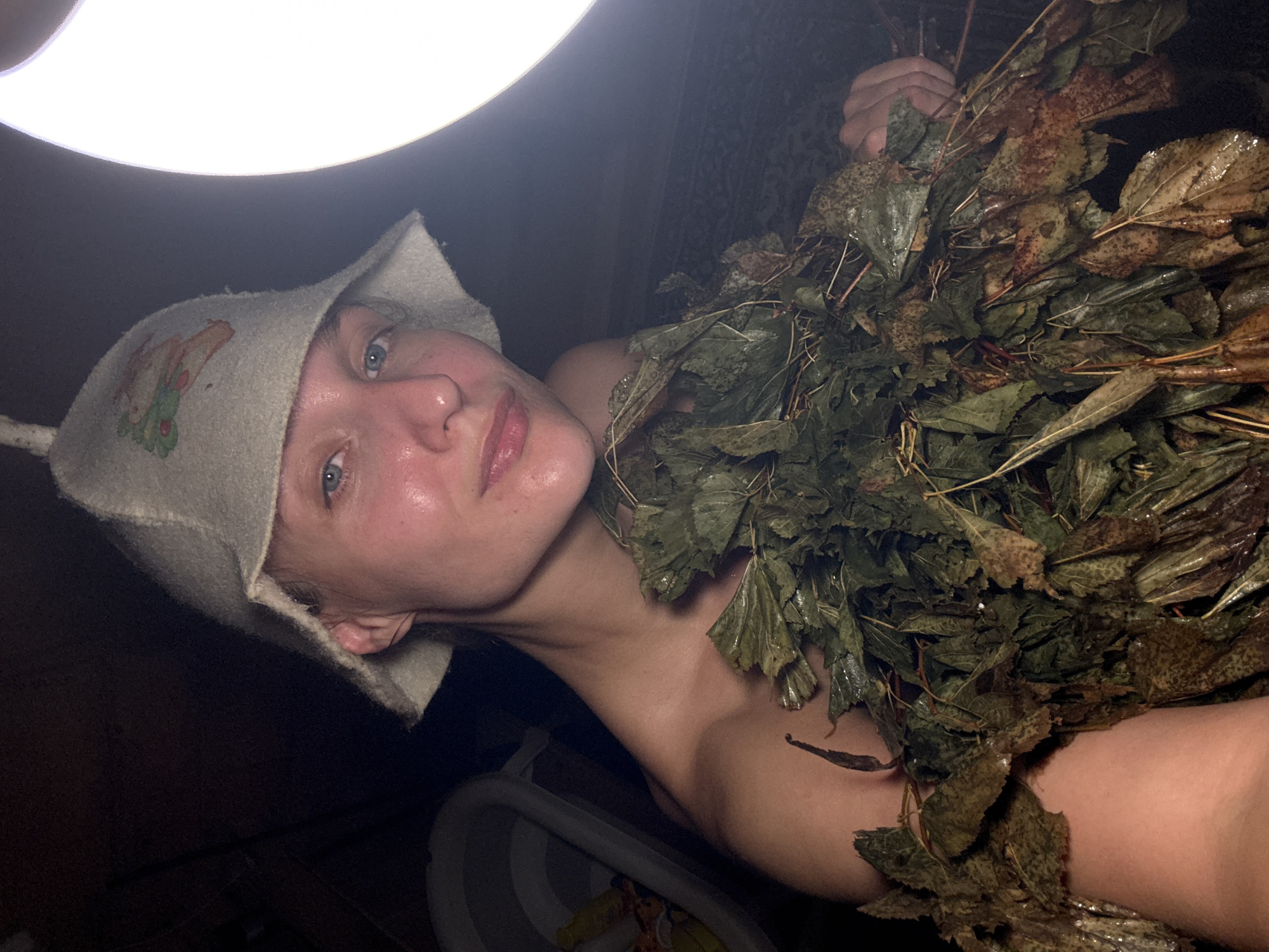 A young woman with blue eyes and fair skin is pictured from the chest up, wearing a light-colored felt hat with an embroidered design. She is holding a bundle of wet, dried leaves and branches, possibly a venik used in Russian banyas. Her shoulders and upper chest are visible, and her skin appears slightly flushed, suggesting a warm environment. She is in a dimly lit room, possibly a banya or sauna. To the left, a portion of a light-colored plastic basin is visible. A bright light source is prominent on the right side of the frame, casting a strong glow. The overall impression is of a private, relaxing moment, likely in a traditional bathhouse setting. The location is identified as Sharkan, Russia.