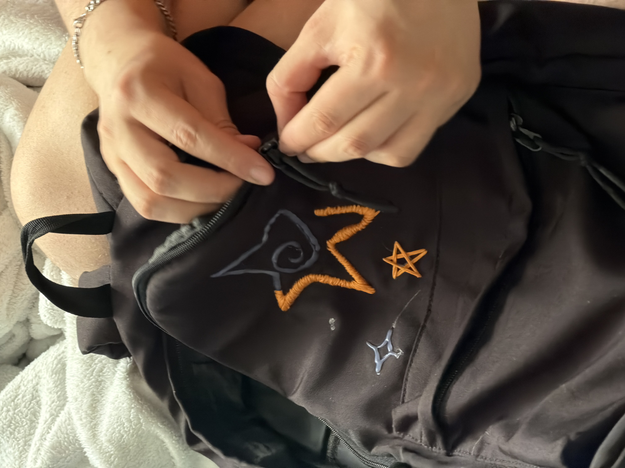 A person is shown closing the zipper of a black backpack. The backpack features embroidered designs, including a star-shaped design with blue and orange thread, a small orange star, and a small blue geometric shape. The person is wearing a silver chain bracelet on their wrist. The backpack is on top of a white blanket. The scene does not show information that allows for identification of its location.