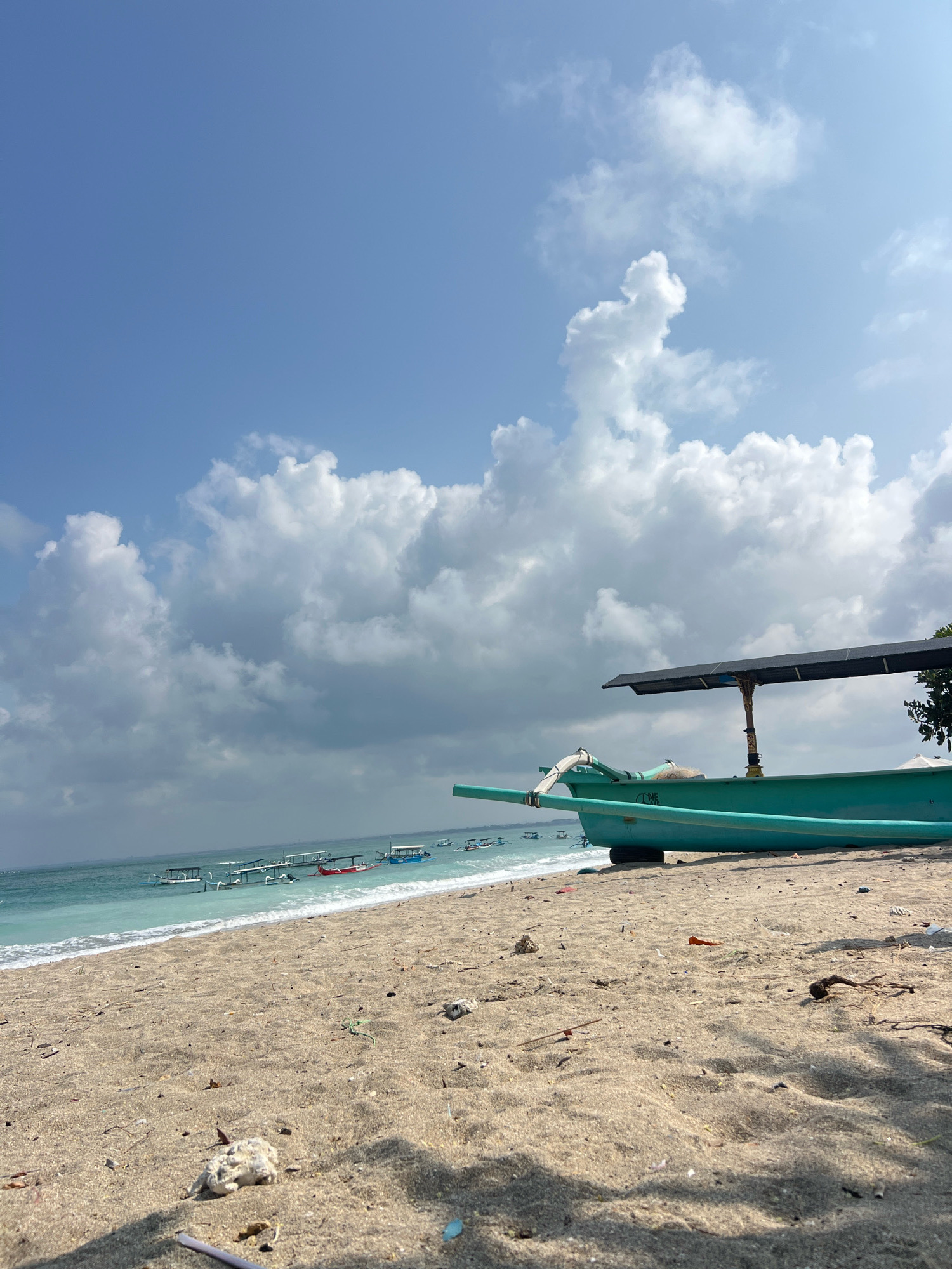 A wide-angle, eye-level shot captures a tranquil beach scene on a sunny day, likely in Kuta, Indonesia. The foreground is dominated by a sandy beach littered with small pieces of coral and debris. In the midground, a vibrant teal-colored boat rests on the shore, its canopy providing shade. Beyond the boat, the turquoise ocean meets the shore with gentle waves. Several other boats are visible further out at sea, some with canopies, suggesting a popular spot for water activities. The sky above is a clear, bright blue, dotted with fluffy white clouds. The scene conveys a sense of peacefulness and the natural beauty of a tropical coastline.
