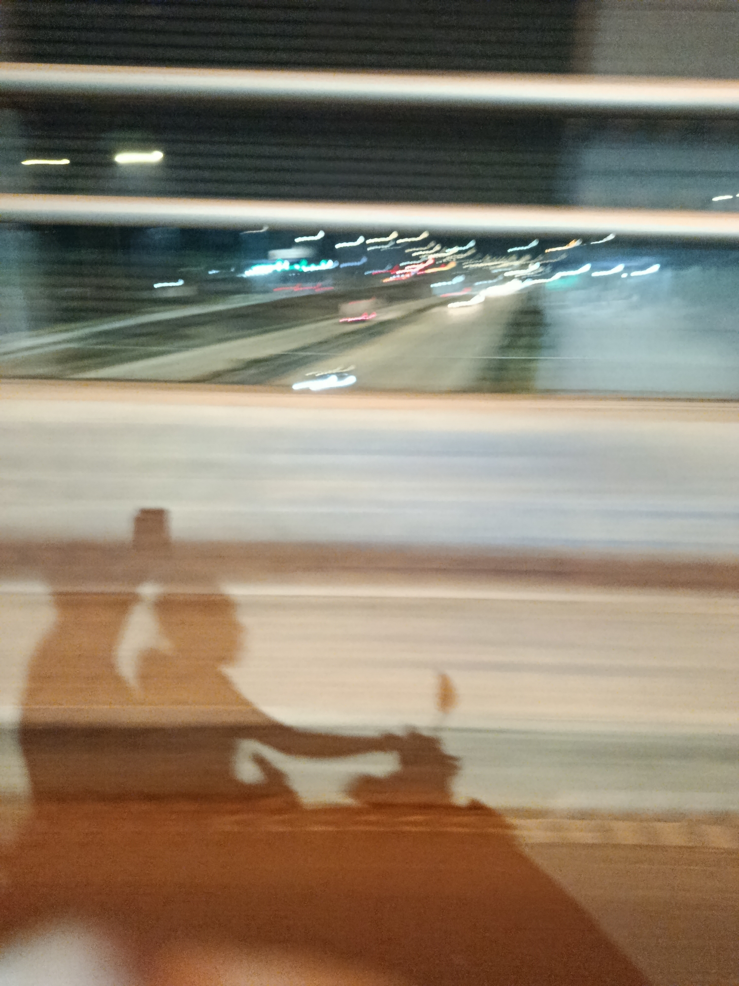 The image depicts a blurred nighttime scene, likely from a moving vehicle. In the foreground, there are distorted shadows suggesting the presence of people and a motorcycle. The shadows are long and elongated, indicating movement and low light conditions. The background shows streaks of light from vehicles on a road, possibly a highway, under a structure that appears to be a bridge or overpass. The overall impression is one of speed and urban night life. The location is Depok, Indonesia.
