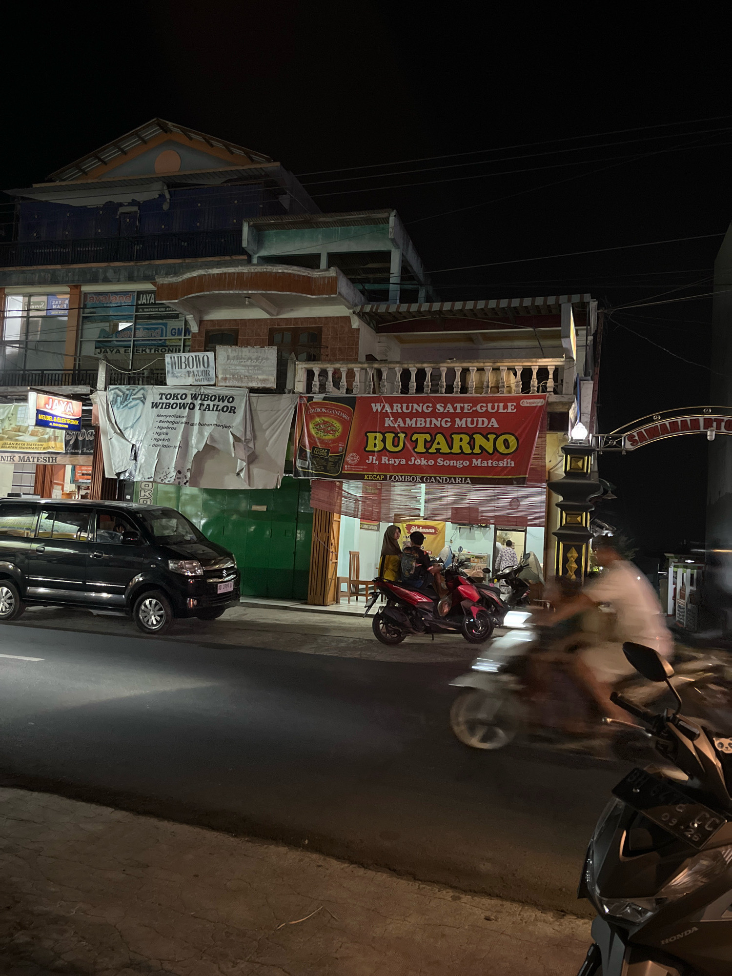 The image captures a nighttime street scene in Matesih, Indonesia. A dark-colored van is parked on the right side of the road, facing left. To its right, a red motorcycle with two occupants is parked in front of a building with red and white striped awnings. Behind them, a large red banner advertises "WARUNG SATE-GULE KAMBING MUDA BU TARNO" and provides an address, "Jl. Raya Joko Songo Matesih." Another banner above it reads "TOKO WIBOWO WIBOWO TAILOR." The building is multi-story with visible windows and balconies. A street lamp illuminates the area. Further to the right, a person on a white motorcycle is blurred due to motion, moving away from the viewer. The road surface appears to be asphalt. The sky is dark, indicating nighttime.