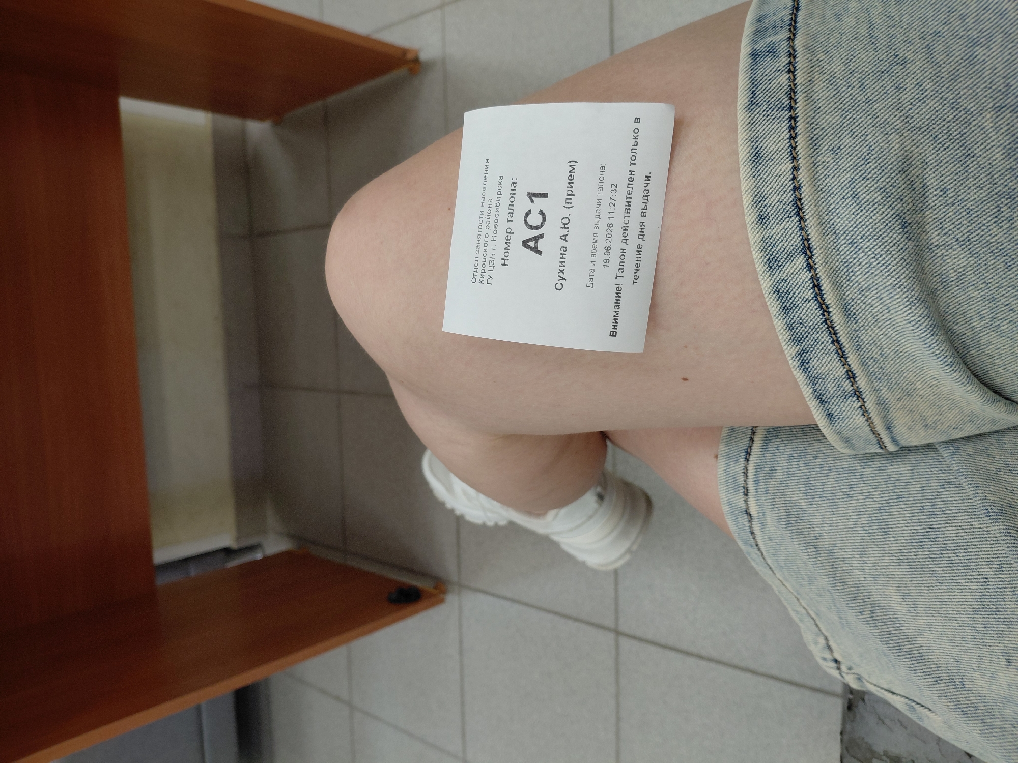 A close-up shot focuses on a person's legs and a paper ticket resting on their thigh. The ticket, a white slip of paper, contains text in Russian. The upper portion of the image shows a wooden desk or table. The floor is tiled with square light gray tiles. The person is wearing a light blue denim skirt and a white sneaker on their left foot. The background is out of focus. The ticket has the following text: "Отдел занятости населения Кировского района ГУ ЦЗН г. Новосибирска Номер талона: АС1 Сухина А.Ю. (прием) Дата и время выдачи талона: 19.06.2026 11:27:32 Внимание! Талон действителен только в течение дня выдачи." This translates to: "Department of Population Employment of the Kirovsky District of the State Employment Service of Novosibirsk. Ticket Number: AC1 Sukhina A.Yu. (reception) Date and time of ticket issuance: 19.06.2026 11:27:32 Attention! The ticket is valid only on the day of issuance."