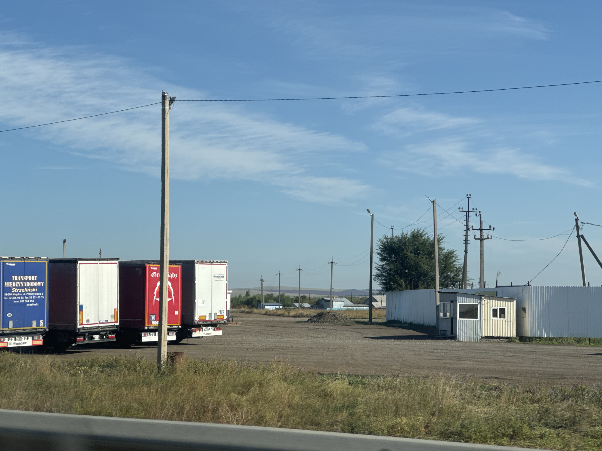 The image shows an outdoor scene in Gamaleevka-1, Russia, on a clear, sunny day with a few wispy clouds in the sky. In the foreground, there is a gravelly lot with several semi-trailer trucks parked in a row. The trucks have various markings, including "TRANSPORT MIĘDZYNARODOWY Strzelinski" on one, and "KRONE" branding on others. The environment is somewhat rural, with tall grass and wildflowers growing along the edge of the lot. In the middle ground, there is a small, utilitarian building that appears to be a guardhouse or small office, constructed with corrugated metal siding and windows. A tall white corrugated metal fence runs behind the building. Further in the background, there are several utility poles with electrical wires, and some scattered buildings and trees suggesting a small settlement or industrial area. The overall impression is one of a logistics or transportation hub in a rural or semi-rural setting.